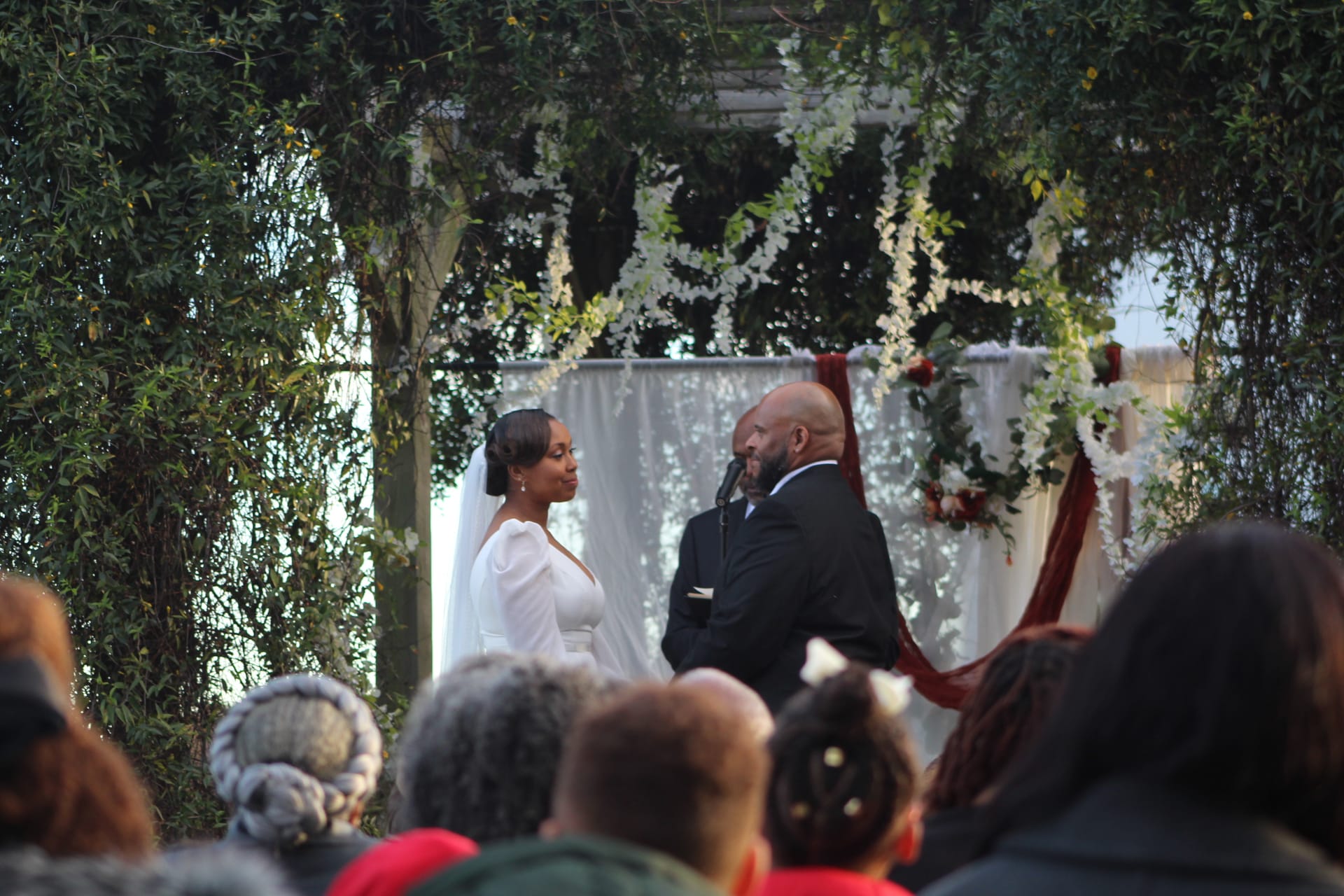 Bride in white gown and groom in black suit exchange vows beneath a lush floral and greenery arch at an outdoor garden wedding ceremony.
