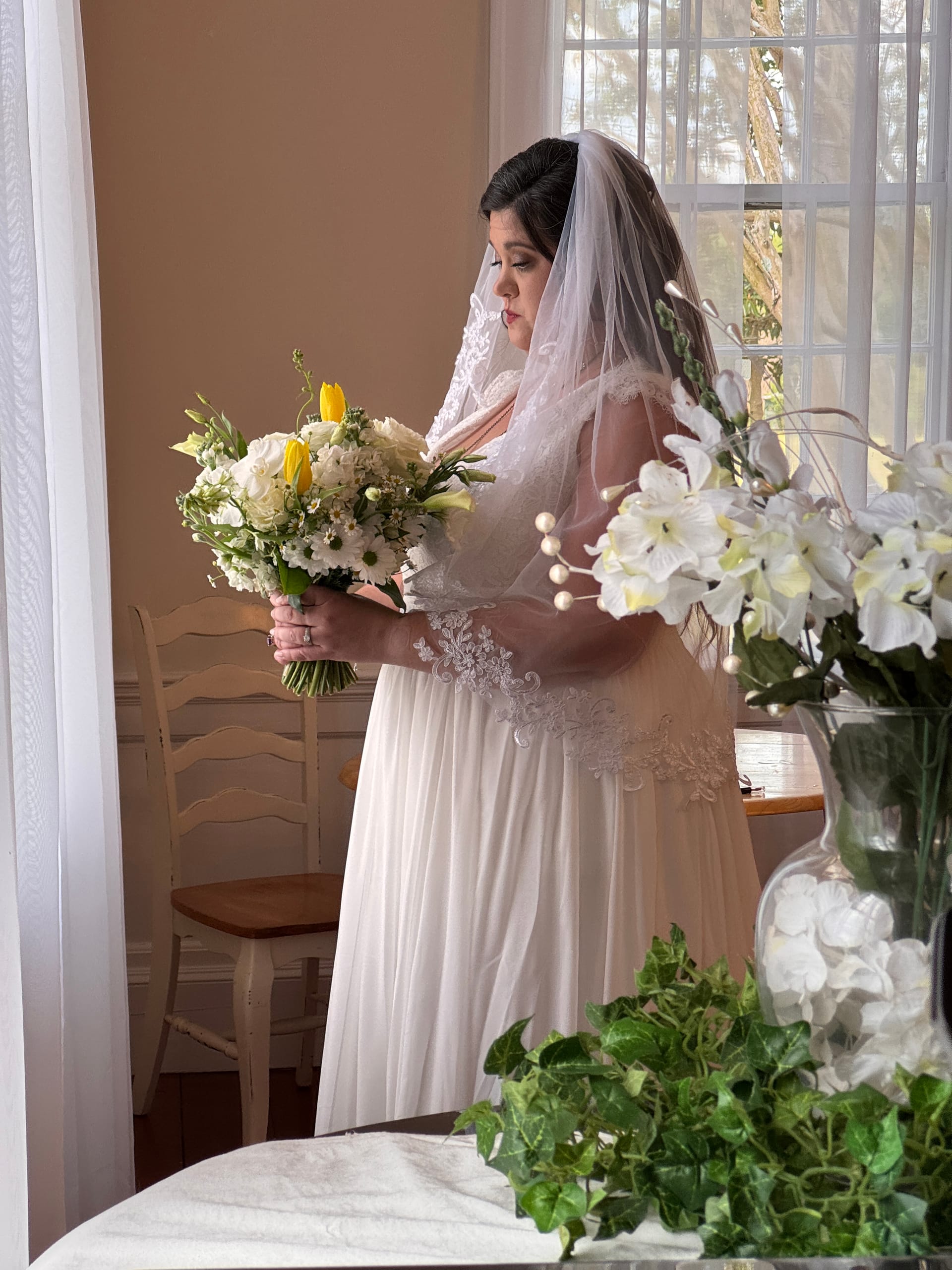 Bride in a lace wedding gown and veil holds a white and yellow floral bouquet while gazing downward near a sunlit window at Scarborough House.