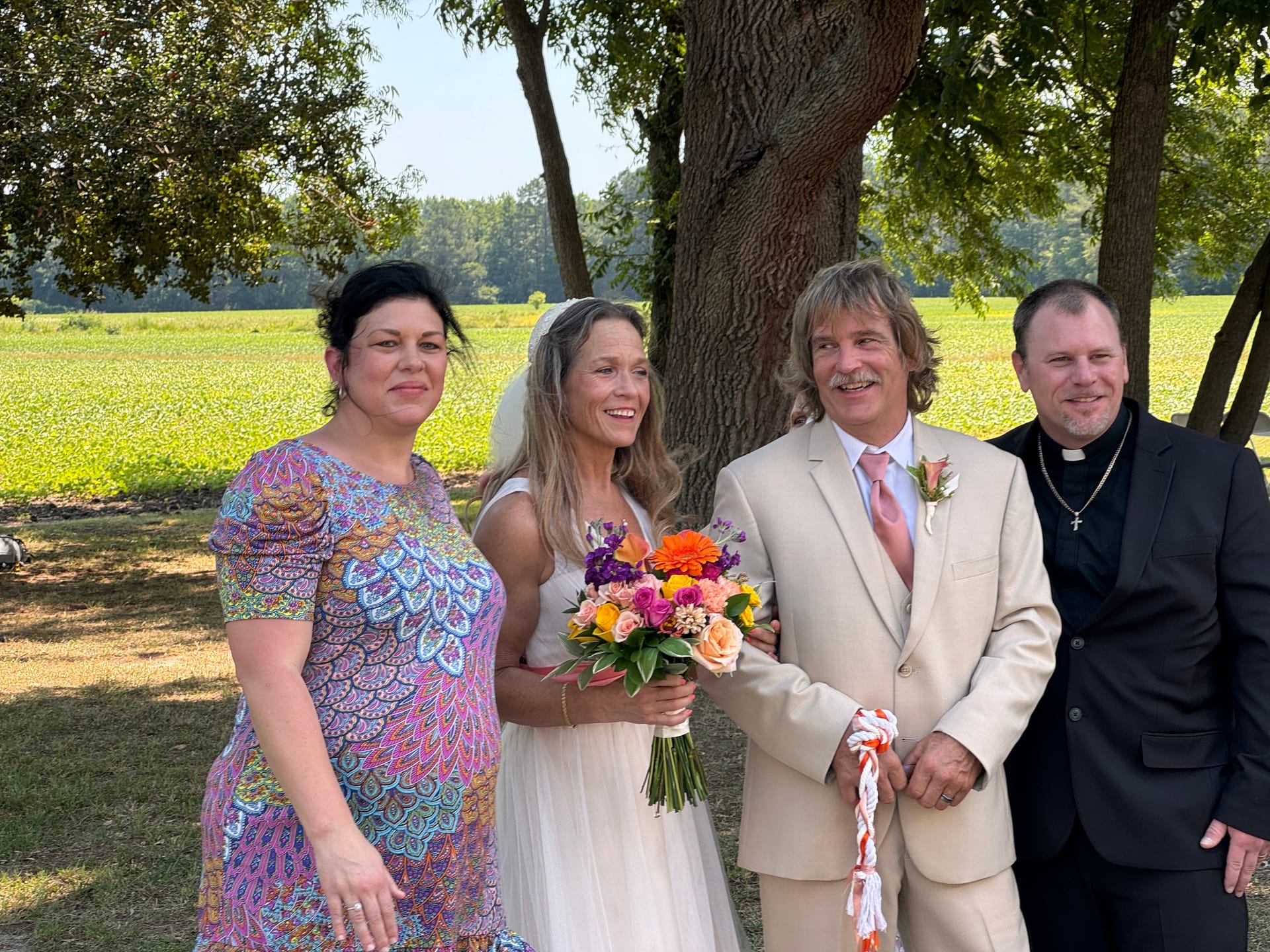Bride holding a colorful bouquet and groom in a tan suit pose with their wedding party officiant and guest beneath a large oak tree overlooking open farmland fields at Scarborough House.