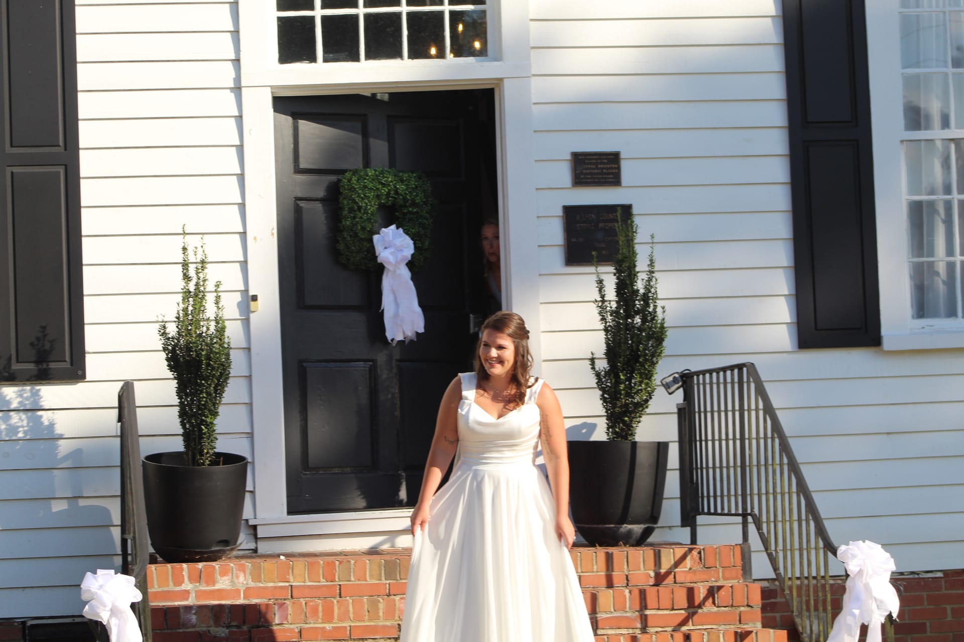 Smiling bride in a white ball gown descending the brick front steps of the historic 1821 Scarborough House manor, flanked by topiary planters and white ribbon bows on the black door.