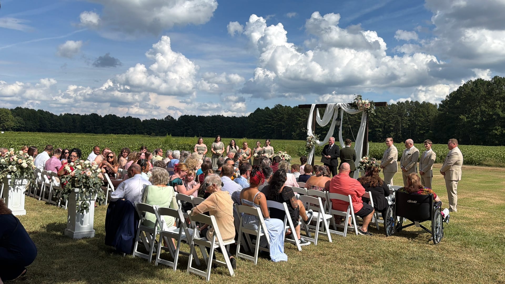 Outdoor wedding ceremony on the lawn at Scarborough House with guests seated in white chairs facing a draped floral arch, wedding party standing alongside, and lush green fields and cloudy blue sky in the background.