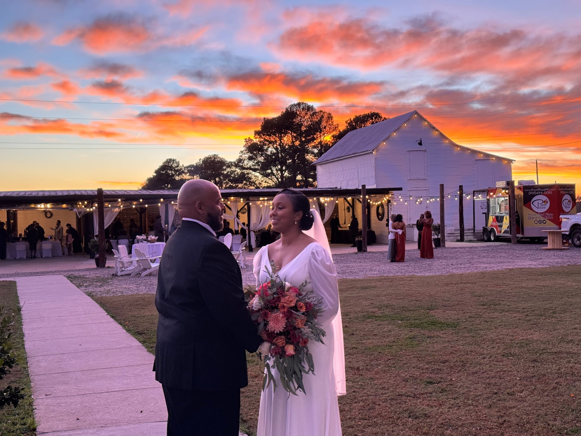 Bride in a flowing white gown holding an autumn bouquet gazes at her groom in a black suit against a dramatic orange and purple sunset sky at Scarborough House wedding venue.