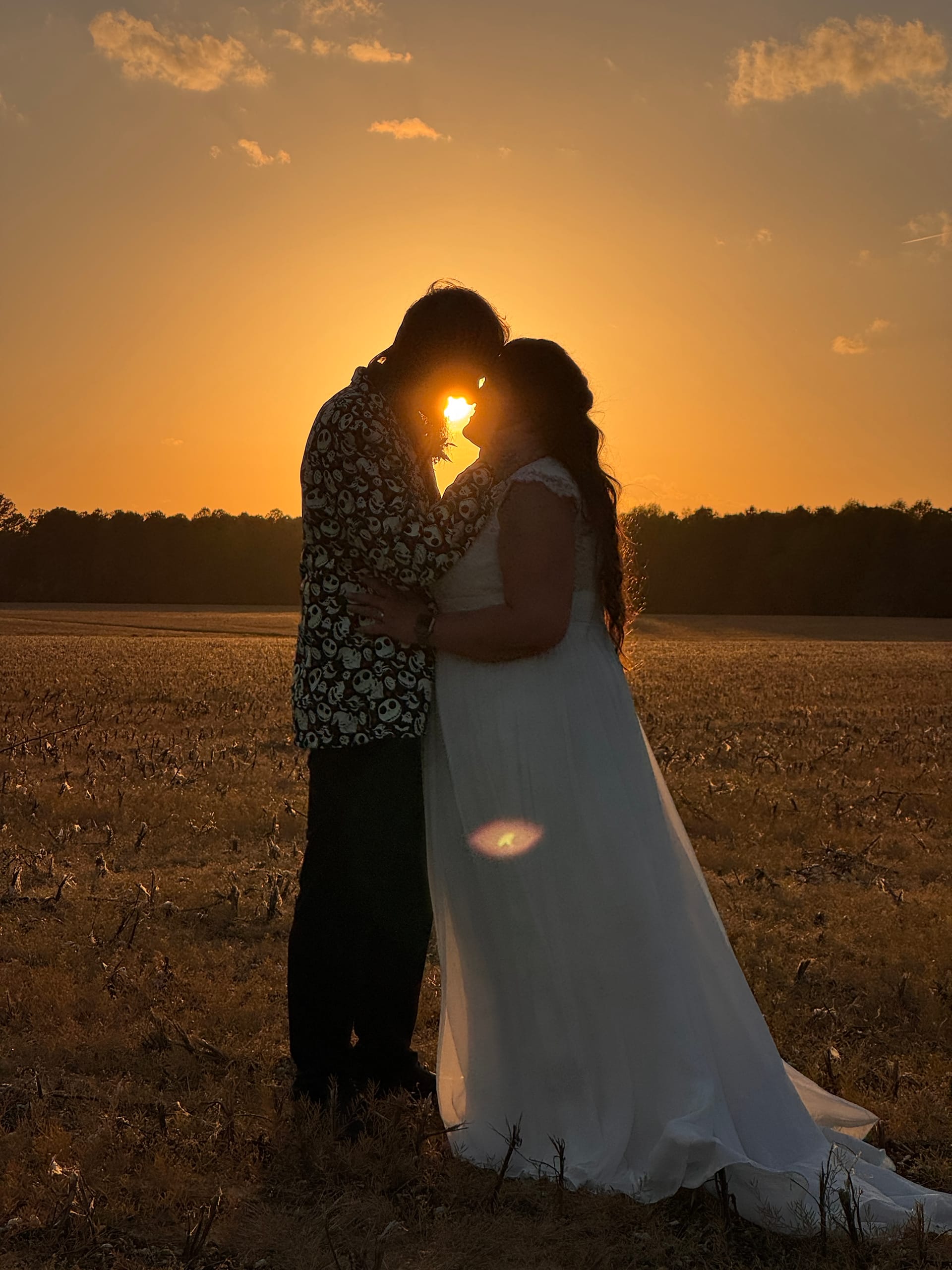 Silhouetted bride and groom embracing at golden sunset over open farmland fields at Scarborough House with the sun glowing between them and the brides long train trailing across the ground.