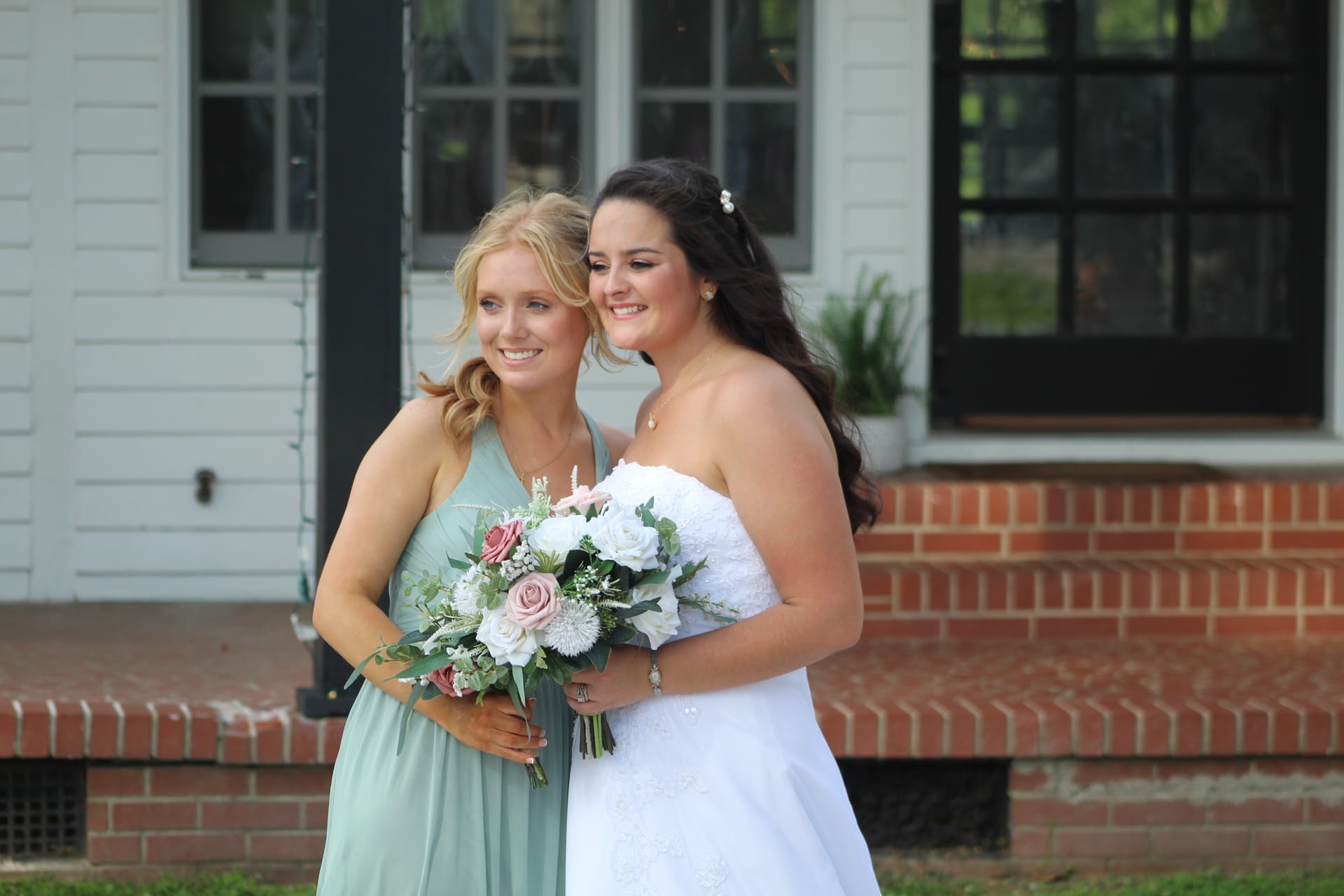 Bride in a white strapless gown and bridesmaid in a sage green dress hold a blush and white floral bouquet while posing together in front of the historic 1821 manor house at Scarborough House.