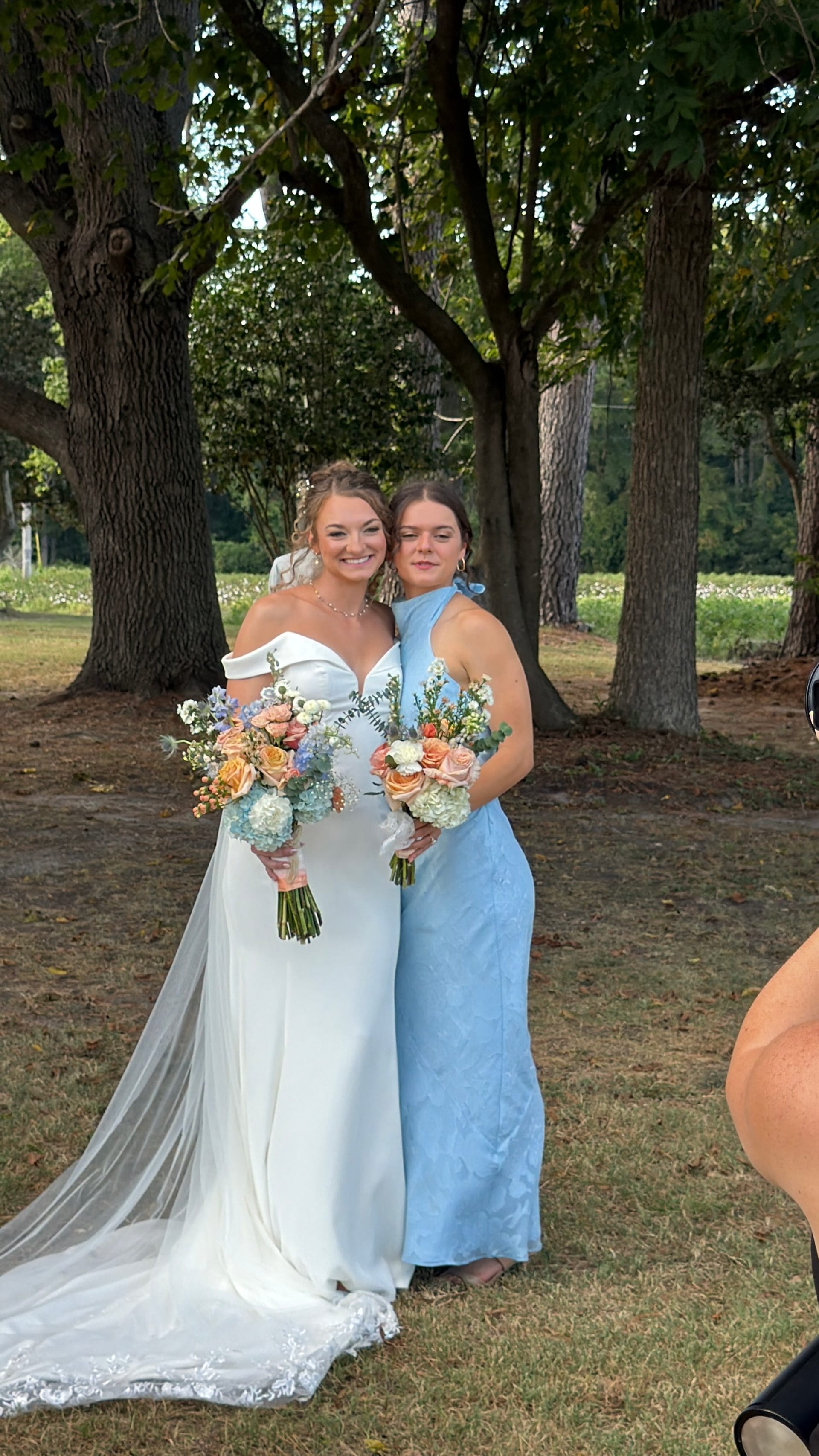 Bride in white off-shoulder gown and bridesmaid in powder blue halter dress smile together holding peach and blue floral bouquets beneath oak trees at Scarborough House estate.