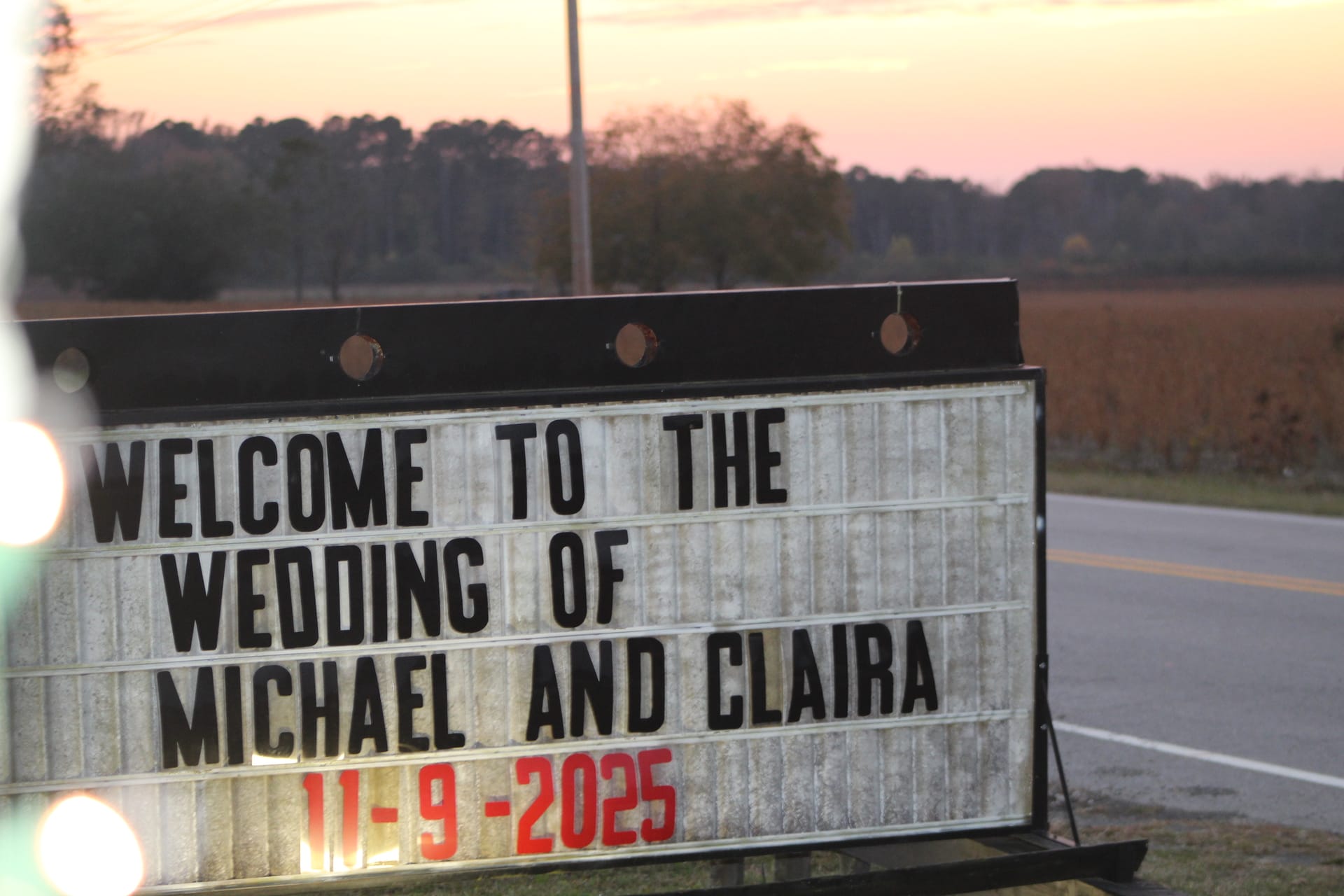 A roadside marquee sign reading Welcome to the Wedding of Michael and Claira 11-9-2025 at dusk with a glowing pink sunset sky and farmland fields in the background.
