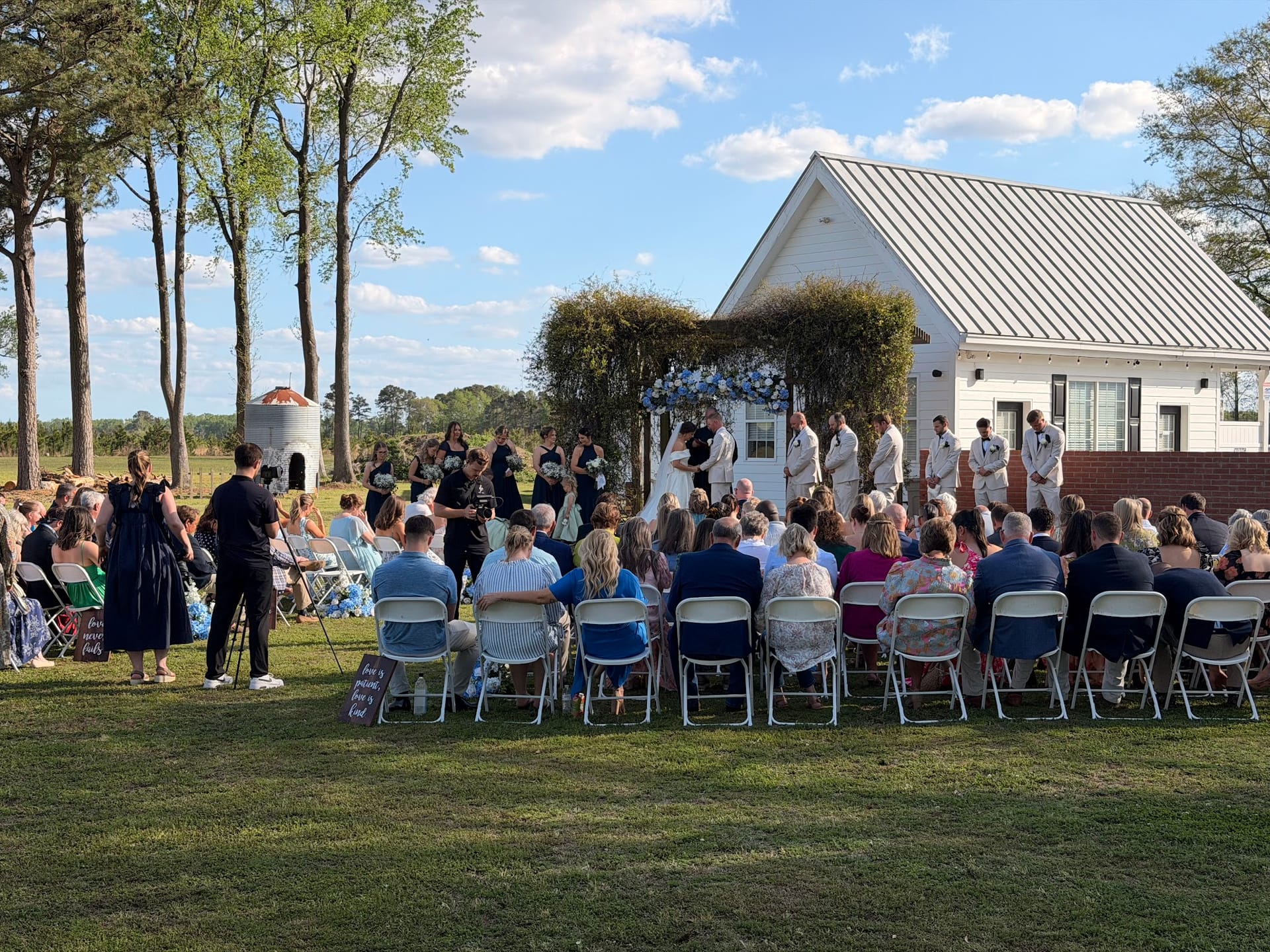 Guests seated in white folding chairs watch an outdoor wedding ceremony beneath a floral arch on the ceremony lawn at Scarborough House historic estate.