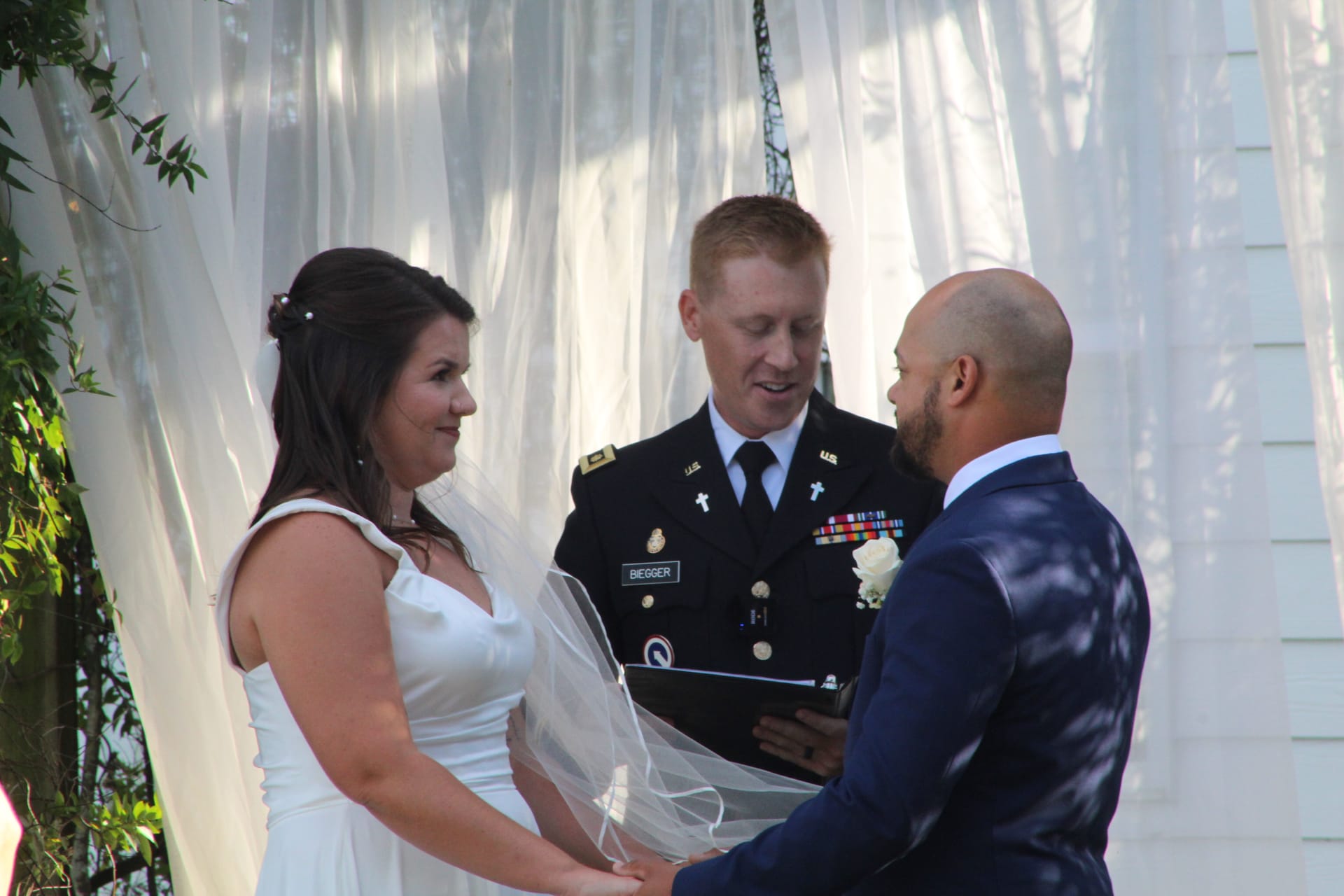 A bride in a white gown and groom in a navy suit hold hands during their wedding ceremony officiated by a US Army chaplain beneath flowing white drapes at a historic estate venue.