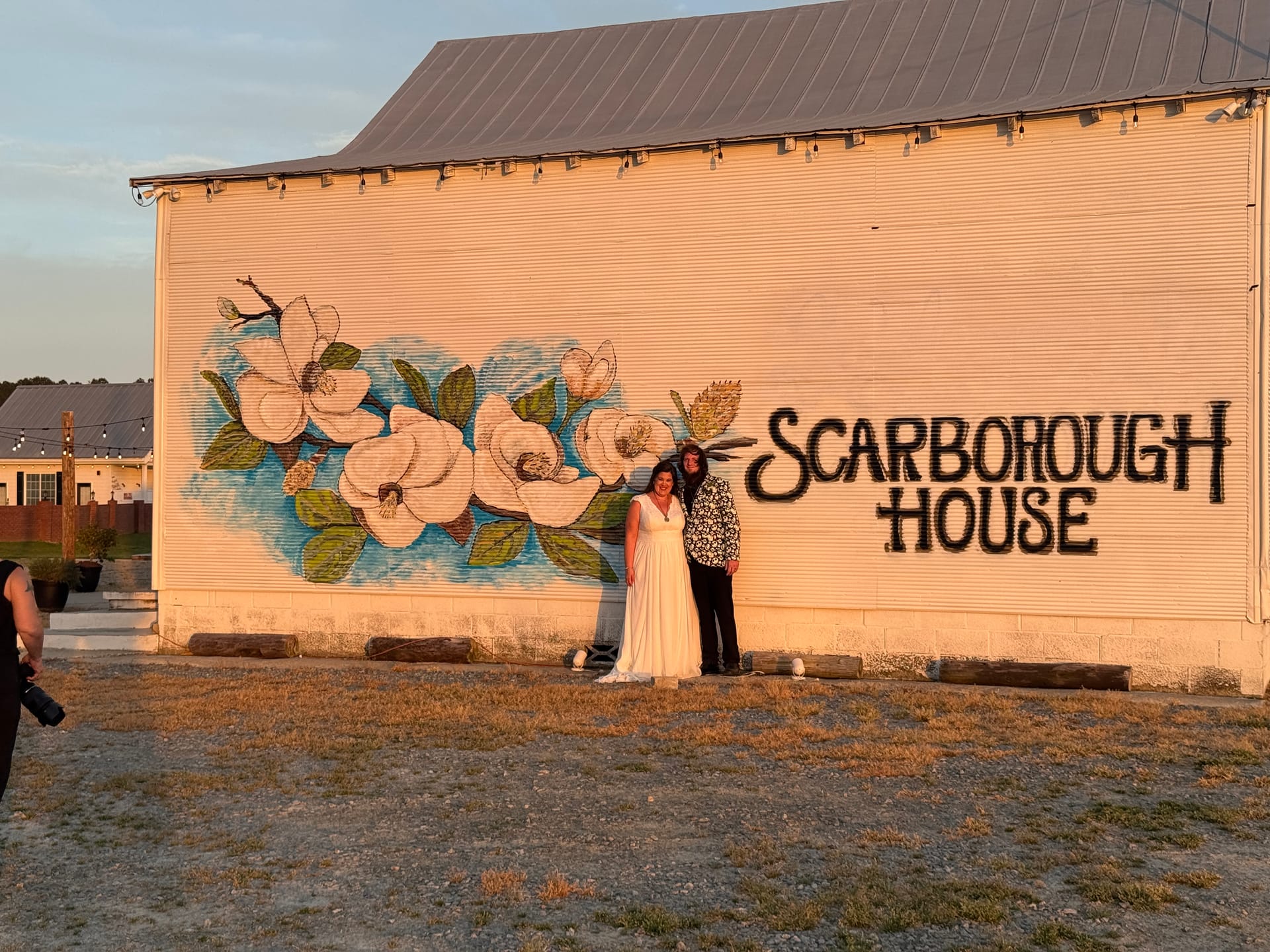 A couple in wedding attire poses together at golden hour in front of the large magnolia flower mural and Scarborough House sign painted on the exterior barn wall of the venue.