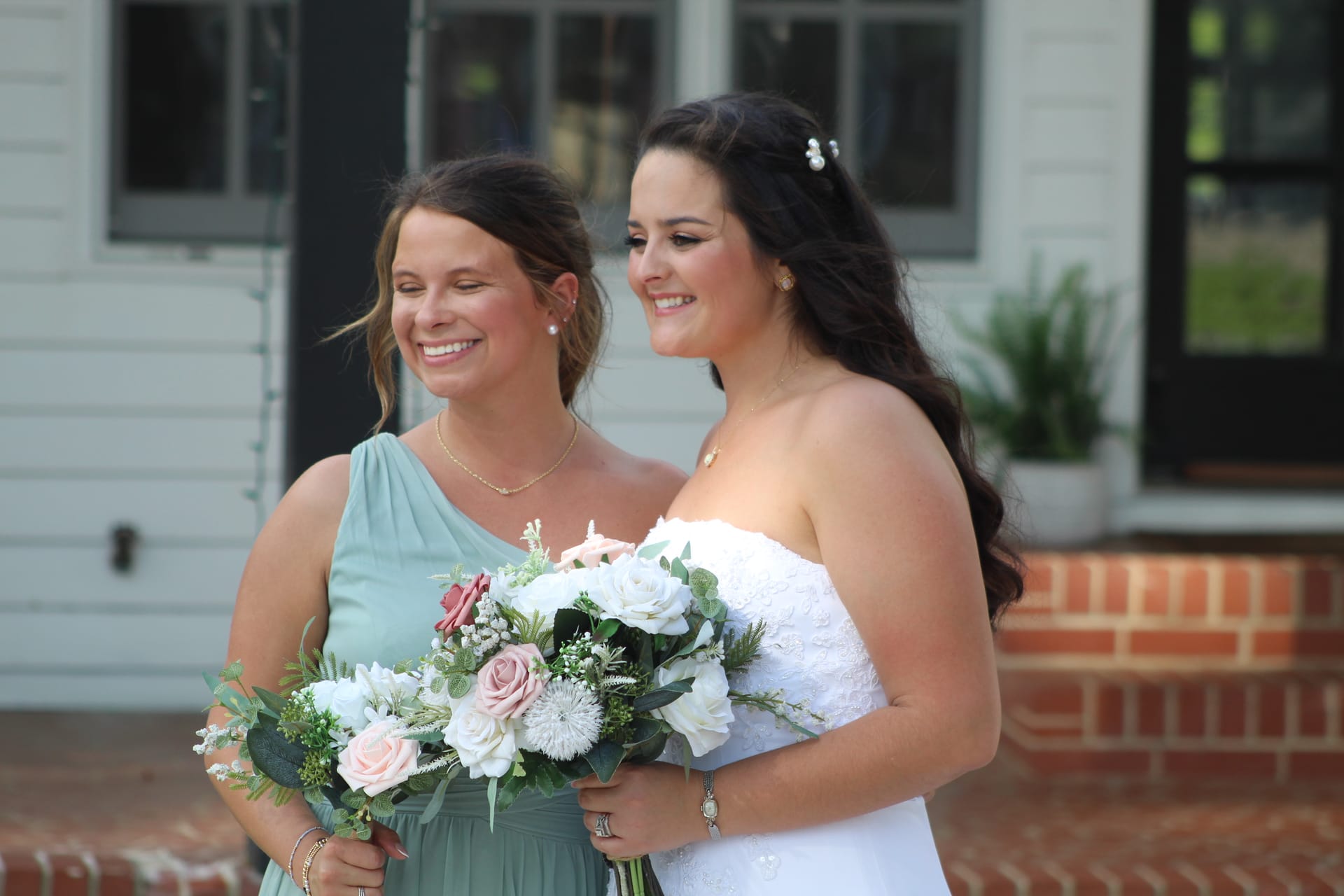 Bride in a strapless white lace gown and bridesmaid in a sage green one-shoulder dress share a joyful moment holding floral bouquets of white and blush roses in front of the historic Scarborough House manor.