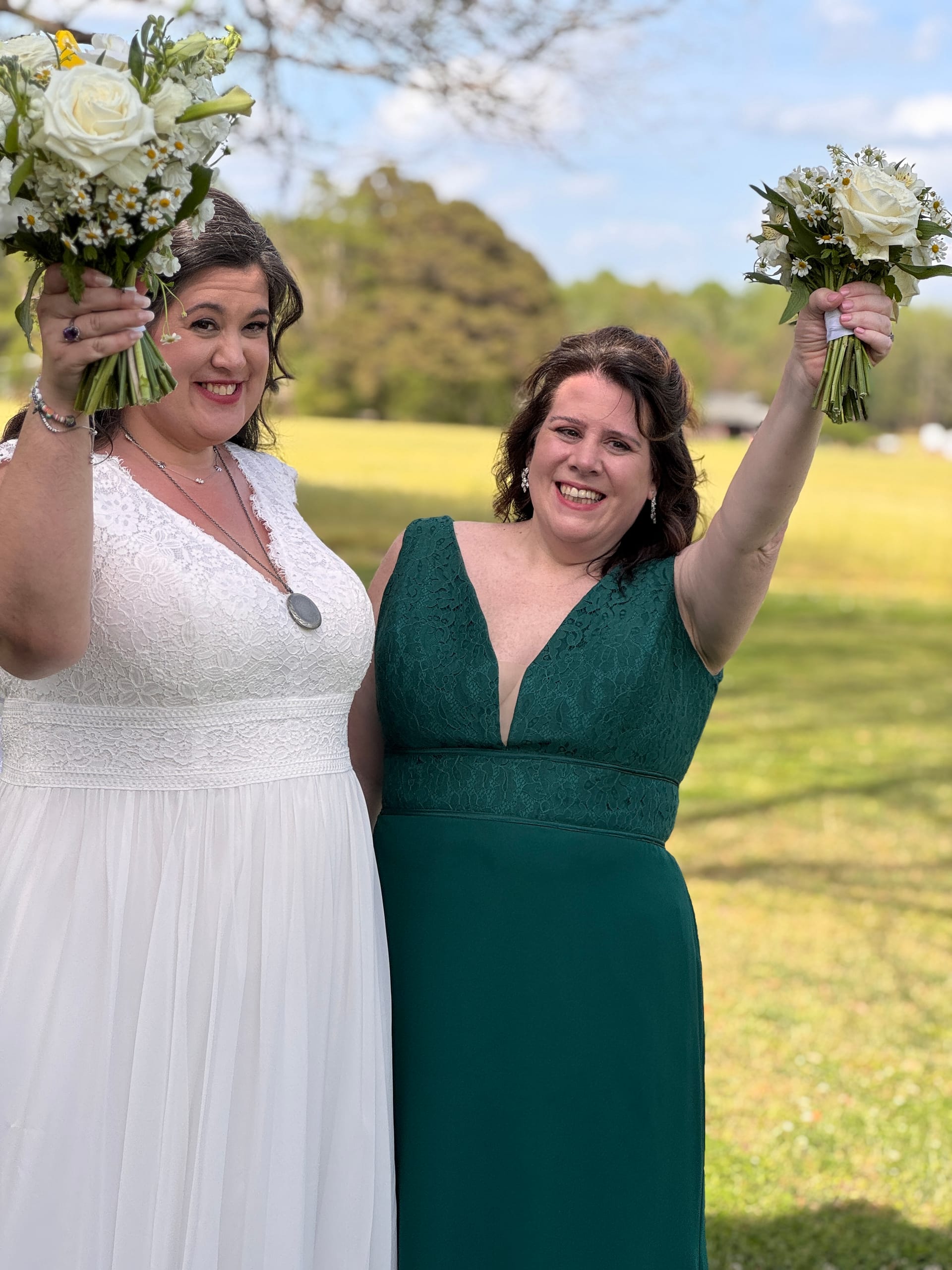 Bride in white lace gown and bridesmaid in emerald green dress joyfully raise their bouquets of white roses and daisies on the sunny ceremony lawn at Scarborough House.
