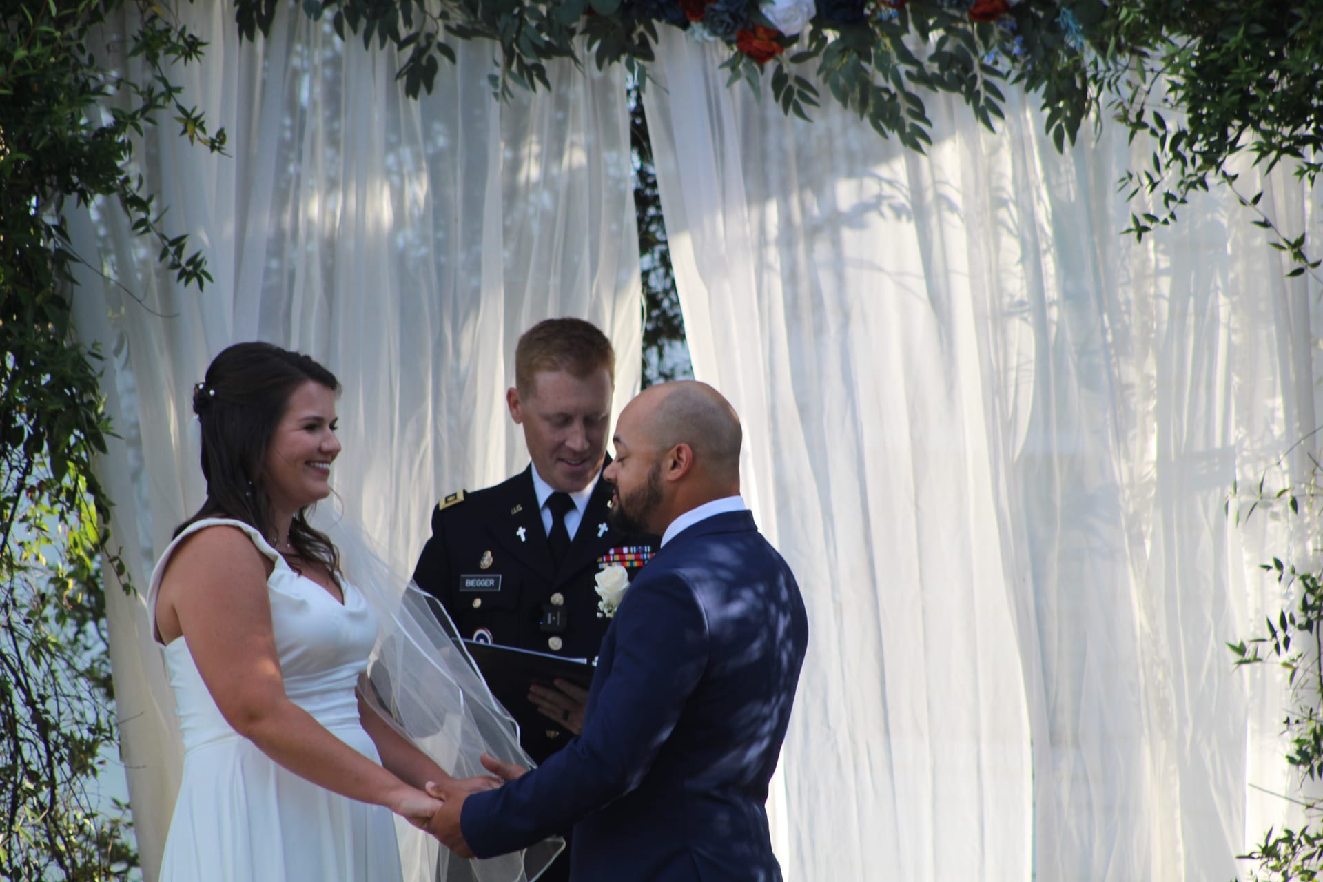 Bride in a white gown smiles joyfully as groom in a navy suit holds her hands during their outdoor wedding ceremony officiated by a military officer beneath a floral and draped arch.