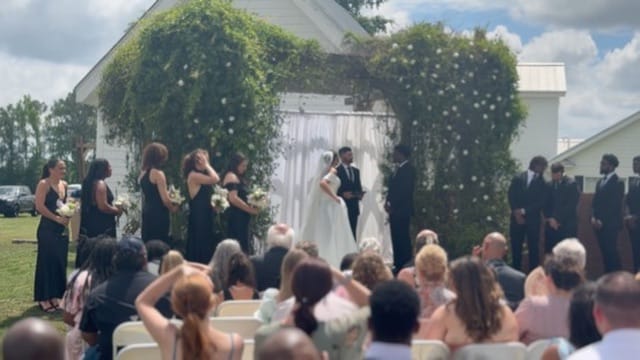Bride and groom exchange vows beneath a lush green floral arch on the ceremony lawn at Scarborough House, surrounded by bridesmaids in black and groomsmen while guests look on.