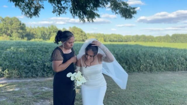 A woman in a black dress helps a bride in a white gown adjust her veil outdoors with green farmland fields and blue sky in the background.