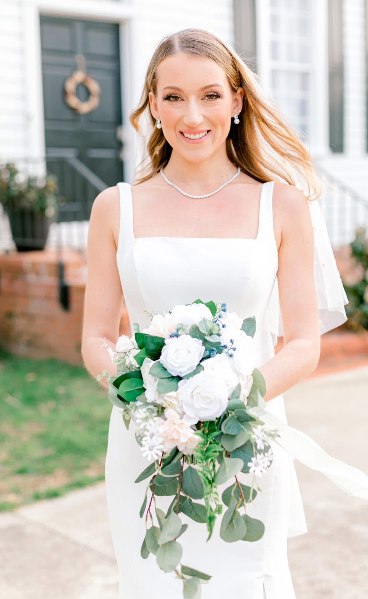 Smiling bride in a white square-neck gown holding a cascading bouquet of white roses and eucalyptus in front of the historic 1821 Scarborough House manor entrance.
