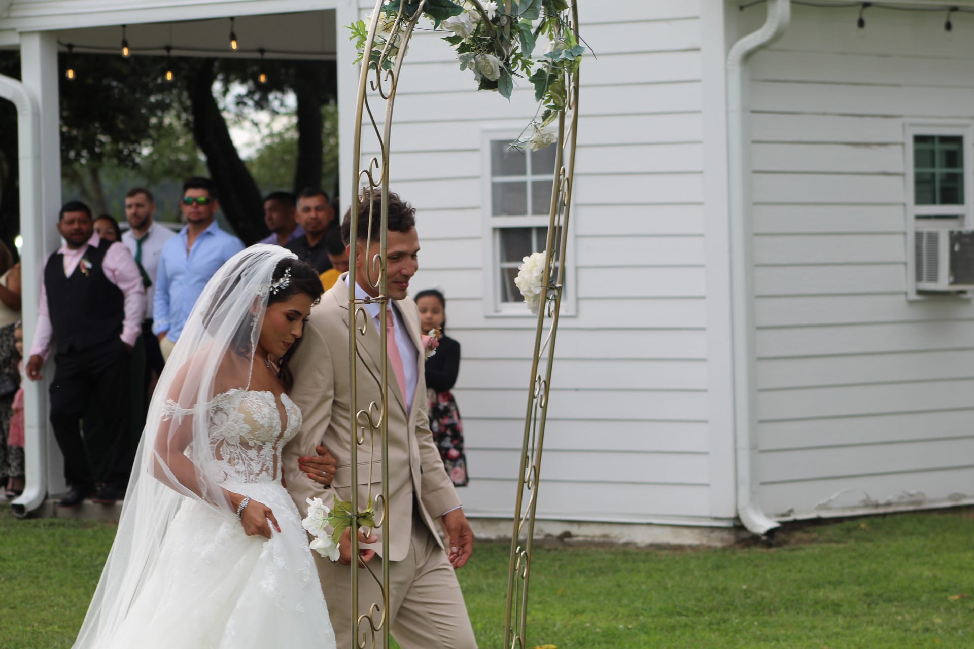 Bride in a lace ballgown and groom in a tan suit walk arm in arm beneath a gold floral arch on the ceremony lawn at Scarborough House historic estate.