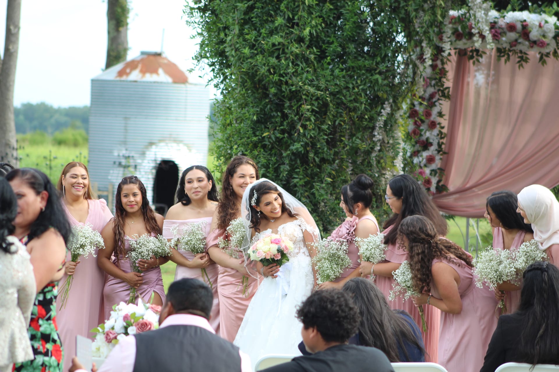 Smiling bride in white lace gown and veil walks down the outdoor ceremony aisle surrounded by bridesmaids in dusty-rose dresses carrying baby's breath bouquets, with lush greenery and a floral-draped arch in the background.