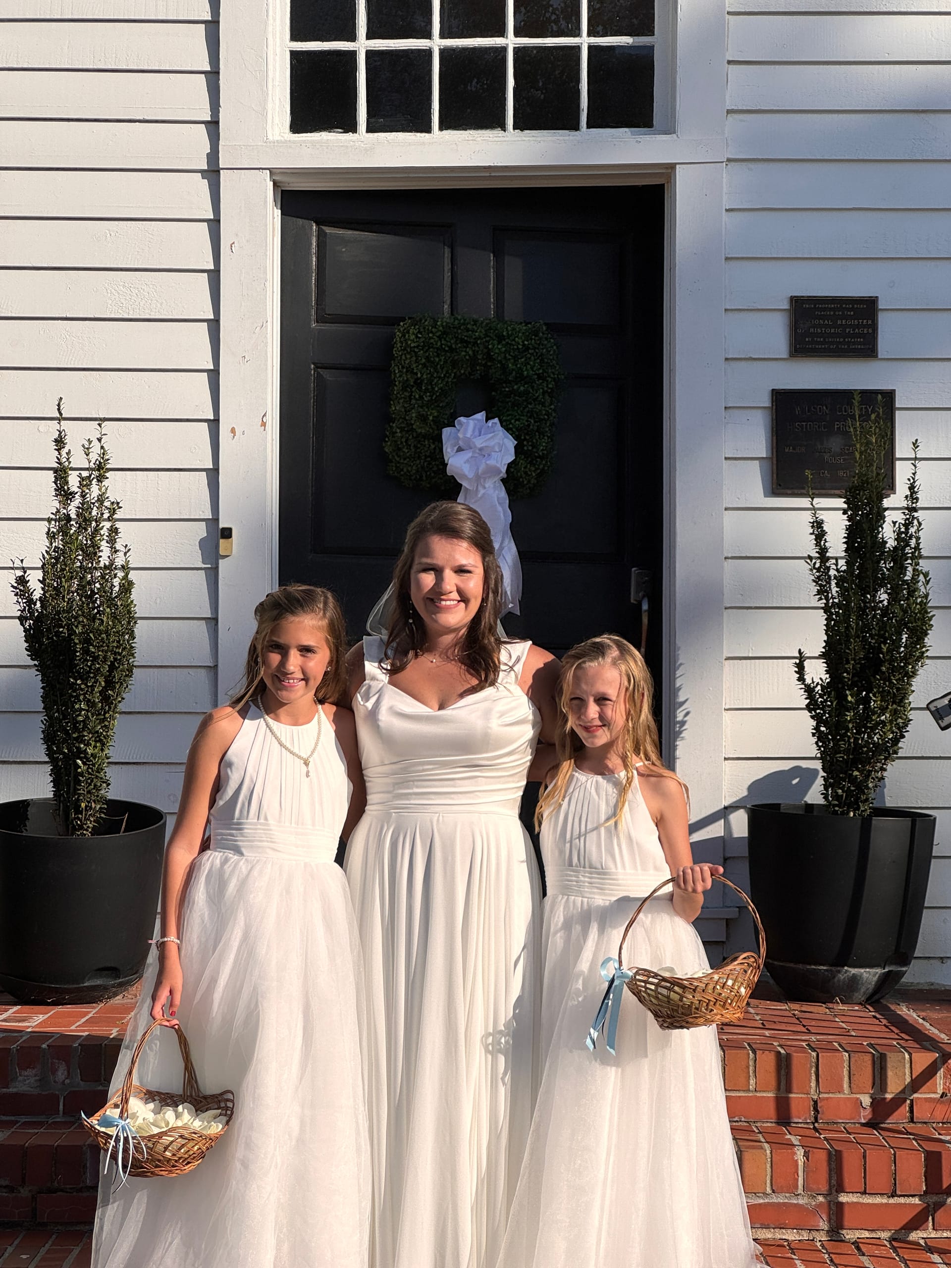 Bride in a flowing white gown poses with two flower girls carrying wicker baskets on the brick front steps of the historic 1821 Scarborough House venue.