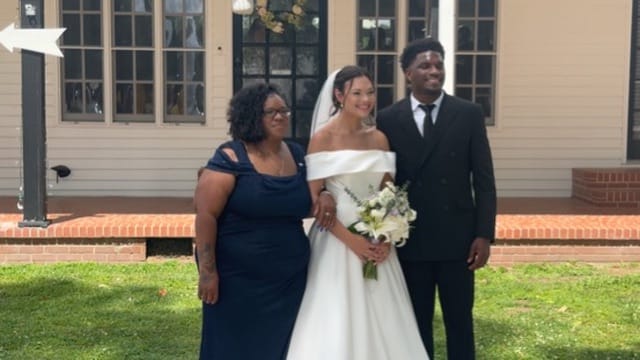 Bride in an off-shoulder white gown holding a white floral bouquet poses with groom in a black suit and a guest in navy blue in front of the historic Scarborough House manor facade.