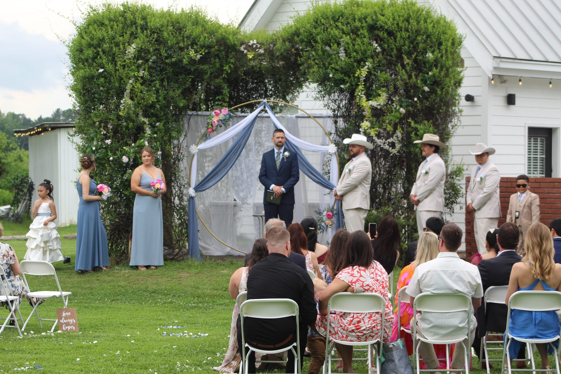 Outdoor wedding ceremony on the Scarborough House lawn with guests seated in white chairs facing the groom and groomsmen standing beneath a circular floral arch draped in dusty blue fabric.