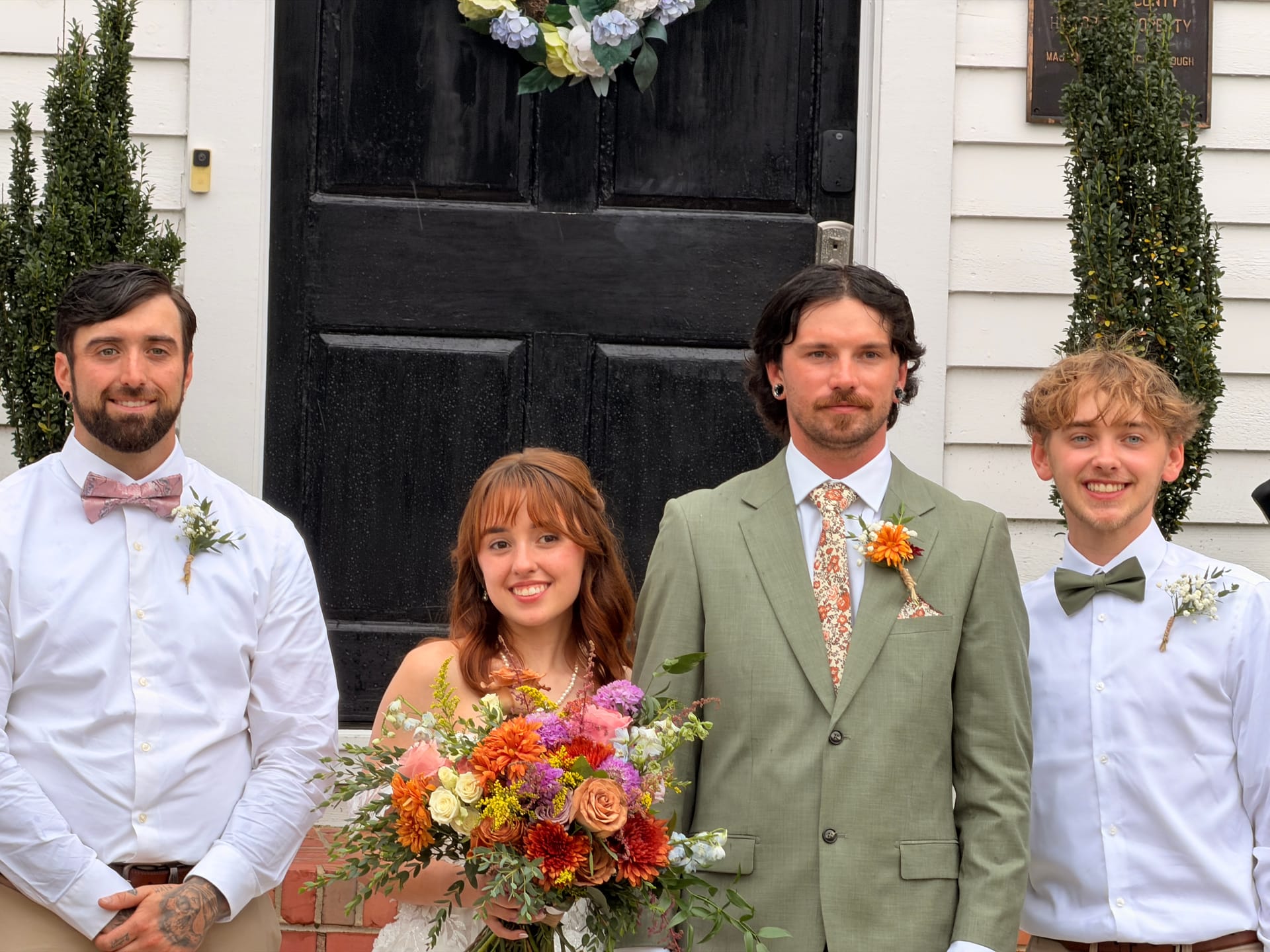 Bride holding a vibrant orange and purple bouquet poses with groom in sage green suit and two groomsmen in white shirts and bow ties on the front steps of the historic 1821 Scarborough House manor.