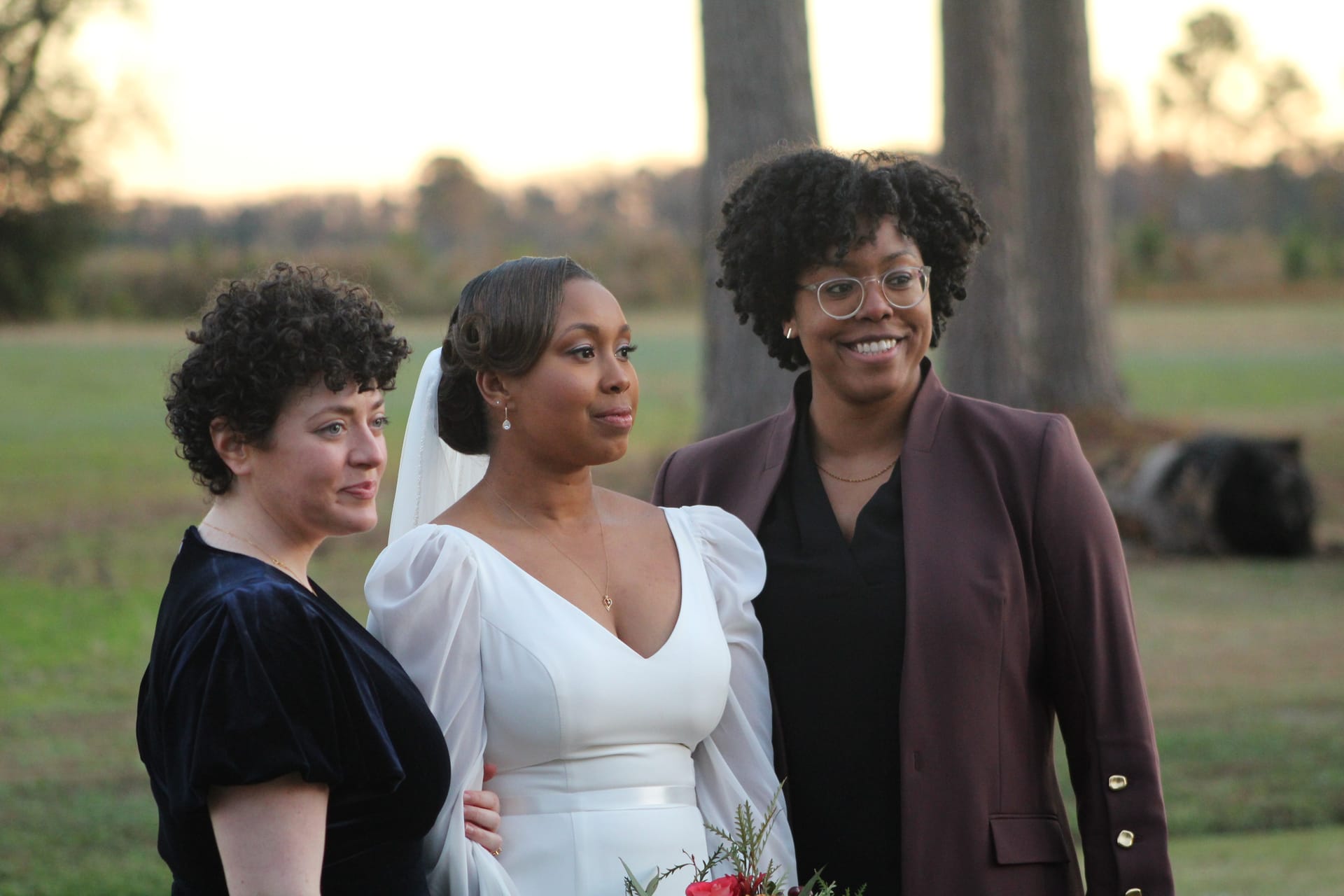 A bride in a white gown and veil poses with two companions in front of open farmland fields at golden hour at Scarborough House estate.