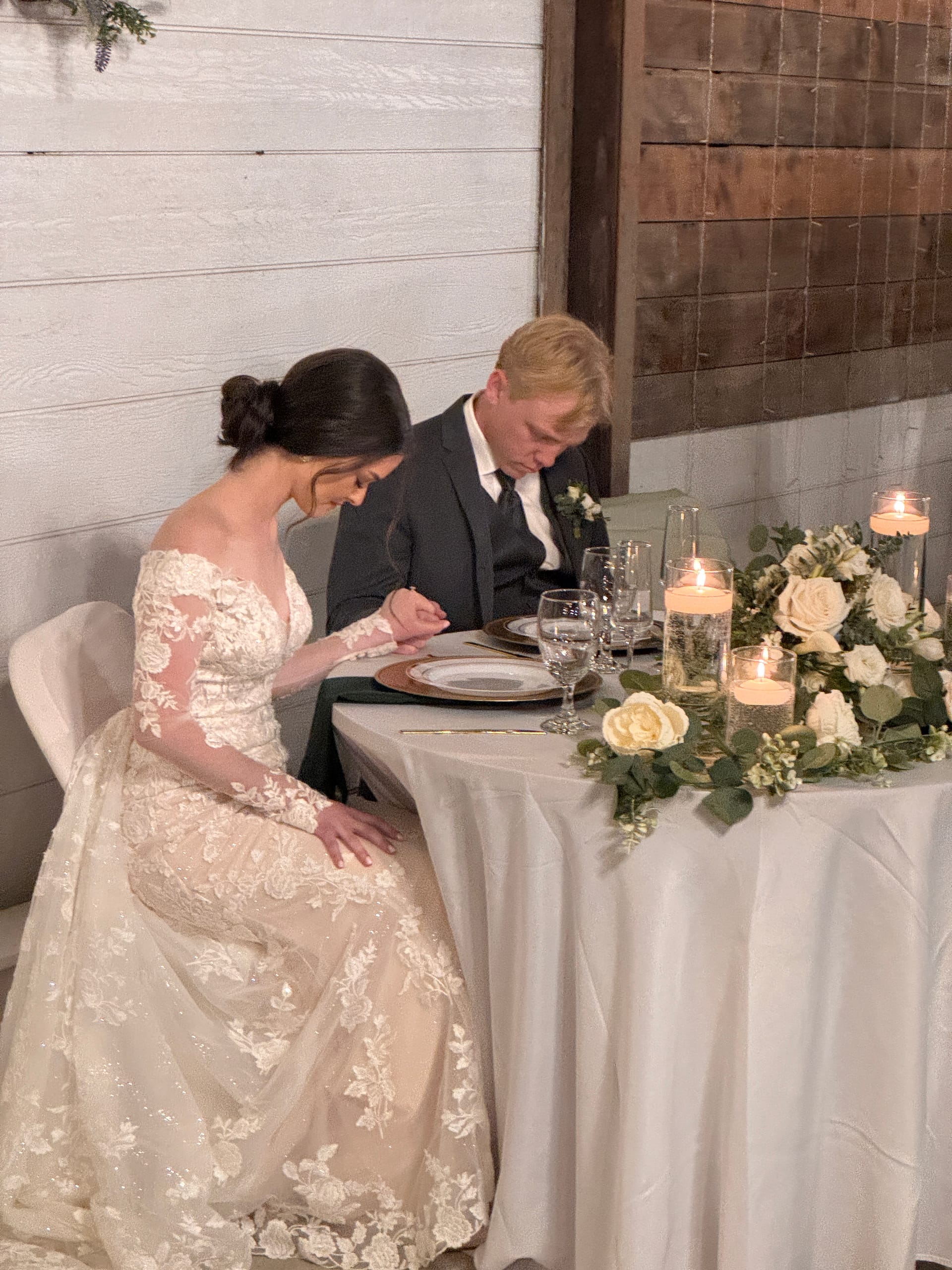 Bride in long-sleeve lace gown and groom in dark suit bow their heads in prayer at a candlelit sweetheart table adorned with white roses and greenery at Scarborough House.