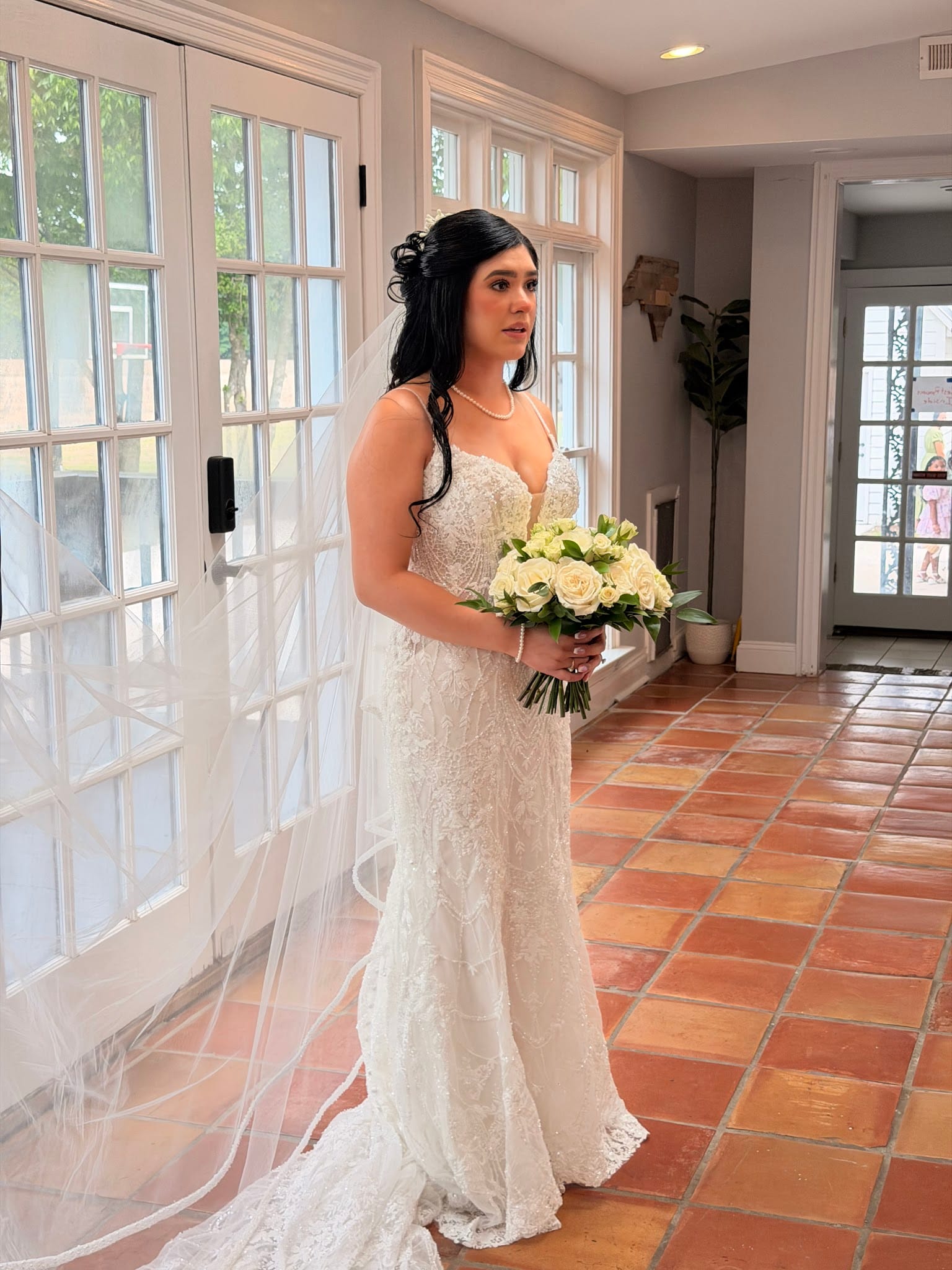Bride in a fitted lace wedding gown holding a white rose bouquet stands near French doors in the grand hall of Scarborough House historic estate venue.