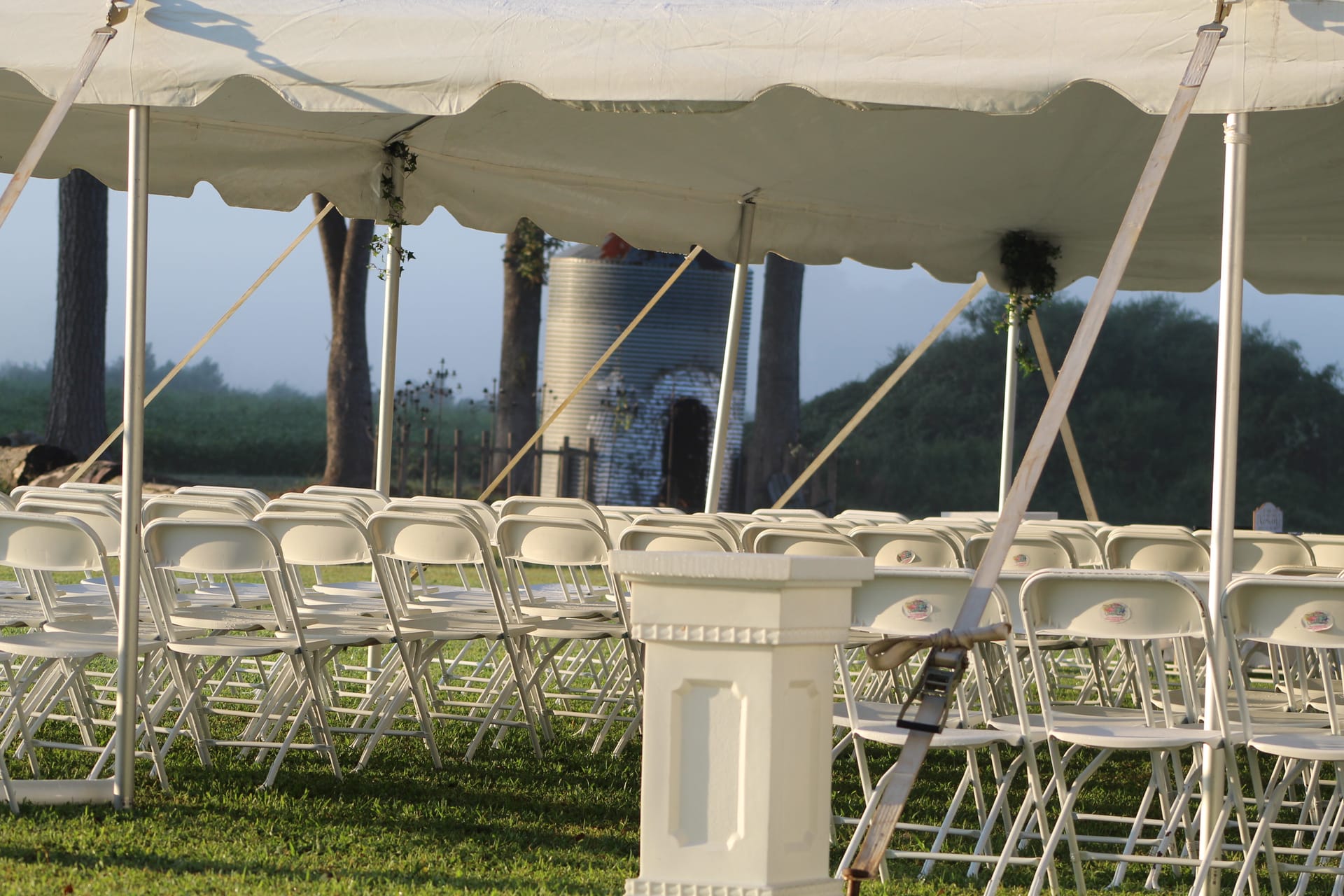 Rows of white folding chairs arranged under a white scalloped tent on the ceremony lawn at Scarborough House, with a rustic grain silo and misty tree line visible in the background.