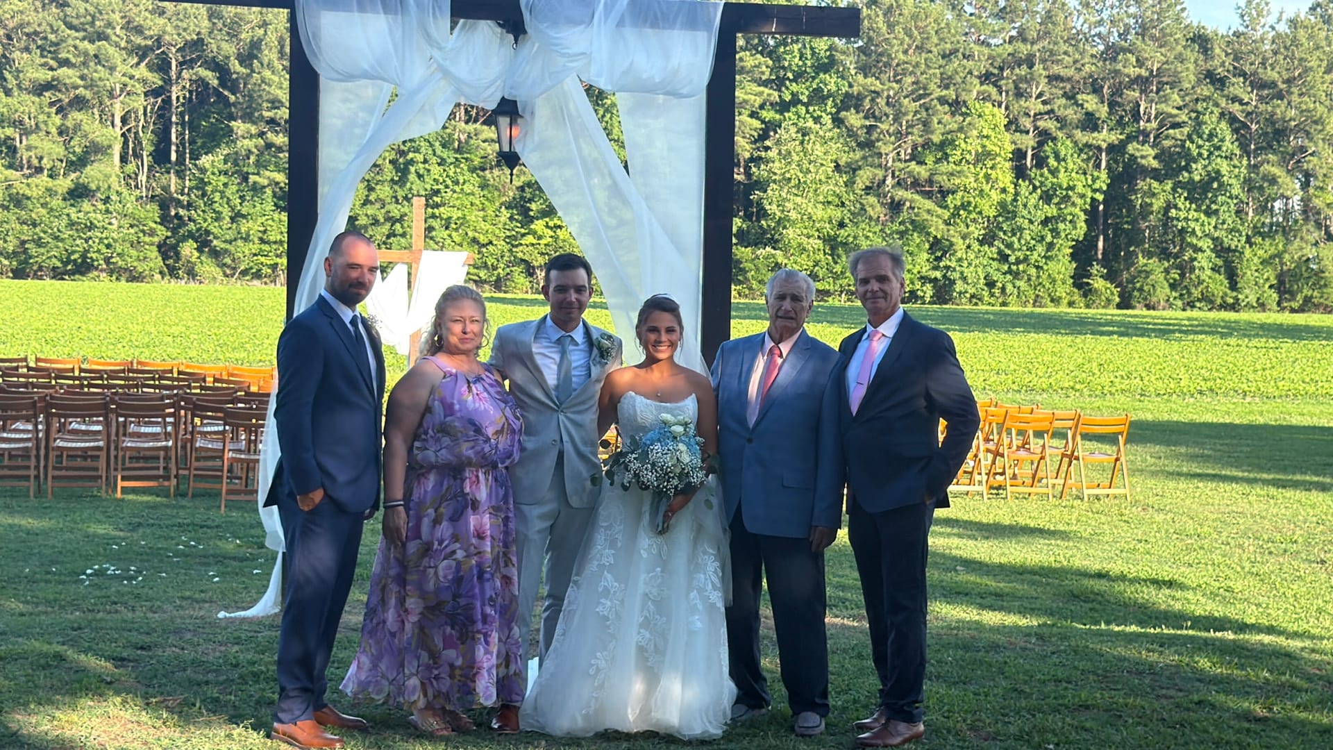Bride and groom pose with four family members under a draped wooden arch on the ceremony lawn at Scarborough House with green fields and trees in the background.