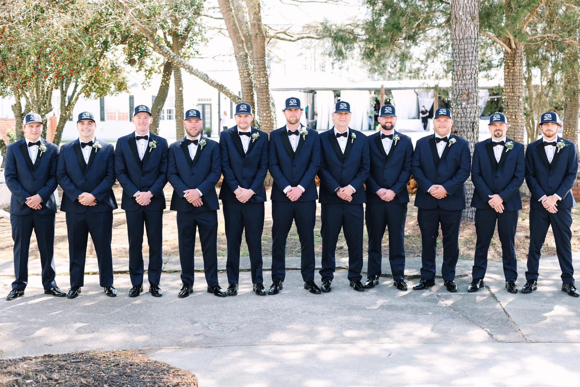 Eleven groomsmen in matching navy tuxedos and black baseball caps stand in a line beneath oak trees on the grounds of Scarborough House historic wedding venue.