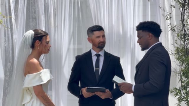 Bride in an off-shoulder white gown and groom in a dark suit exchange vows before an officiant against a flowing white drape backdrop at Scarborough House.