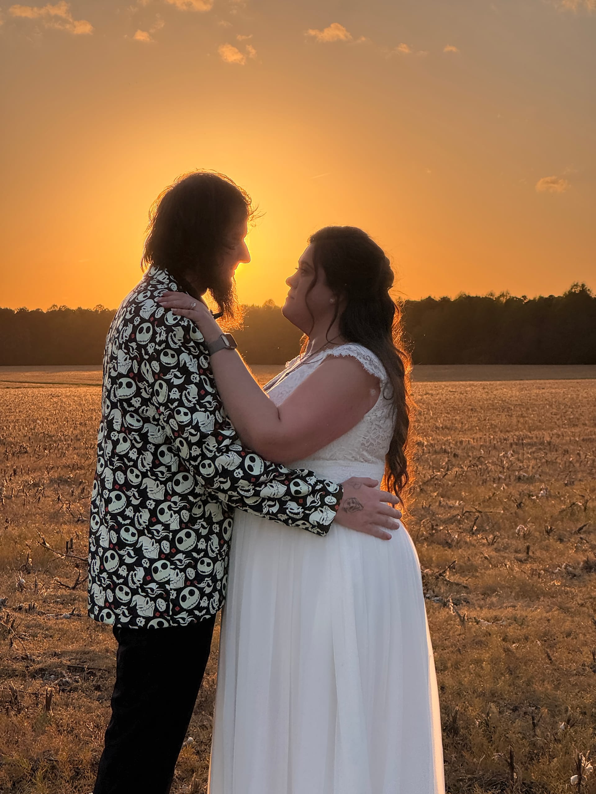 Bride in white lace gown and groom in a Nightmare Before Christmas patterned blazer embrace facing each other in an open field bathed in a vivid golden sunset at Scarborough House estate.