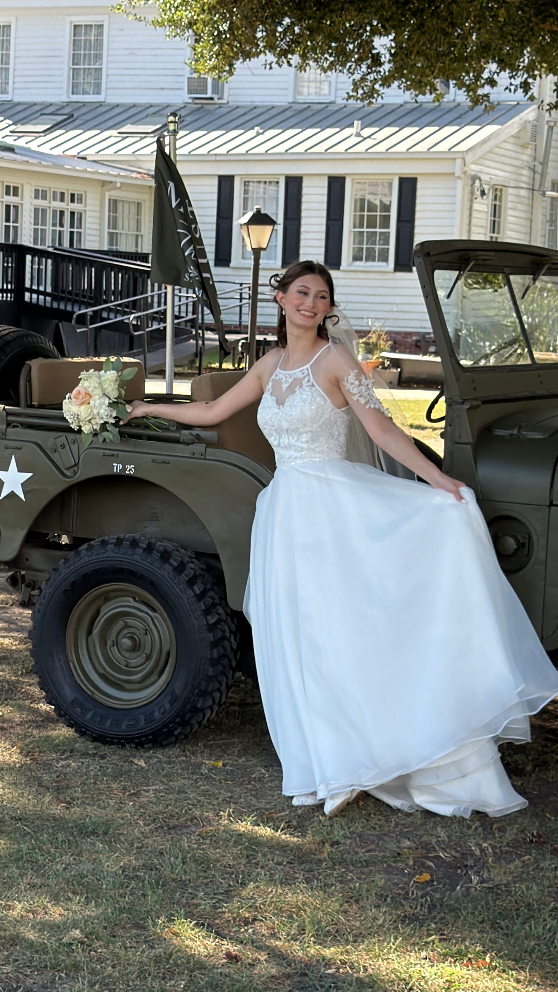 Smiling bride in a white lace gown holding a floral bouquet posed beside a vintage military jeep in front of the historic 1821 Scarborough House manor.