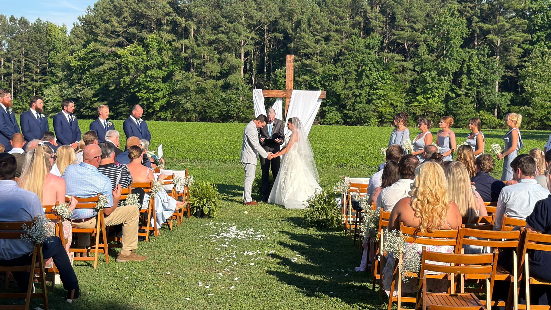 Bride and groom exchange vows before a wooden cross altar draped in white fabric on the ceremony lawn at Scarborough House, surrounded by groomsmen in navy, bridesmaids in blue, and seated guests on a sunny summer day.
