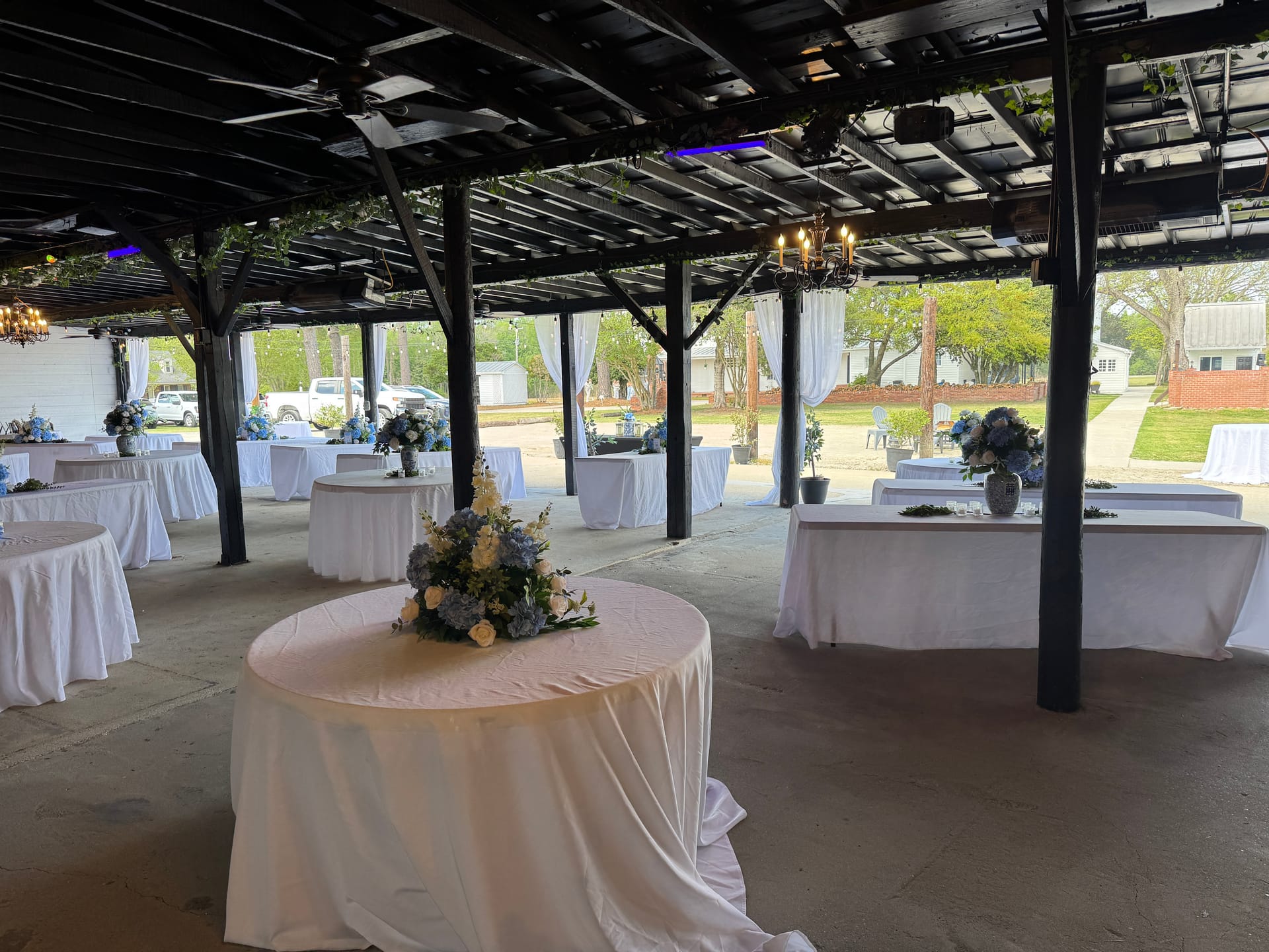 Elegantly set open-air pavilion at Scarborough House featuring round tables draped in white linen and adorned with blue hydrangea and white rose centerpieces beneath a dark wood pergola strung with soft lighting.