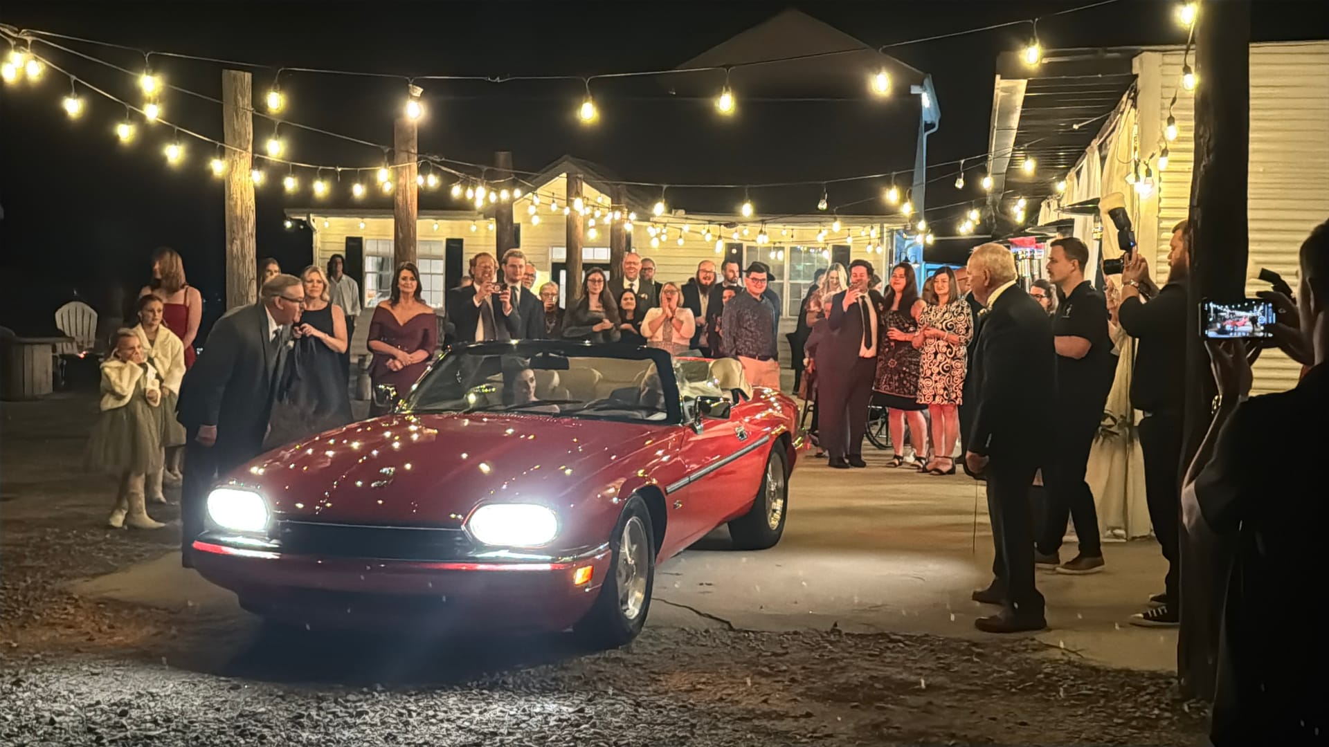 Newlyweds depart in a classic red convertible convertible surrounded by cheering wedding guests under warm festoon string lights at a historic outdoor venue at night.