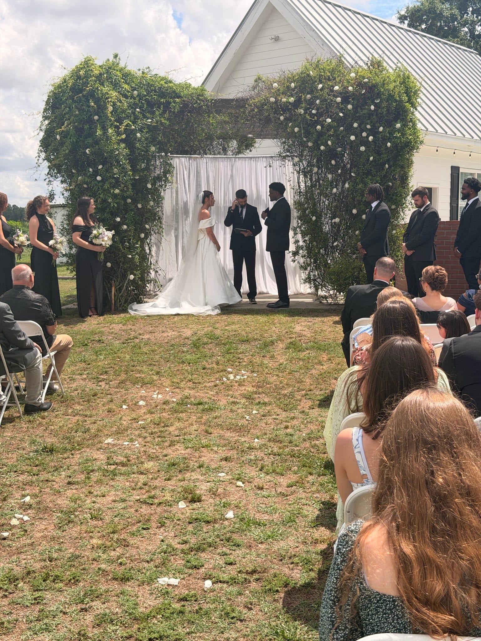 Outdoor wedding ceremony on the lawn at Scarborough House with bride in white gown and groom exchanging vows beneath a lush ivy arch adorned with white florals and draped fabric, surrounded by bridesmaids in black and groomsmen while guests are seated in white chairs along the aisle.