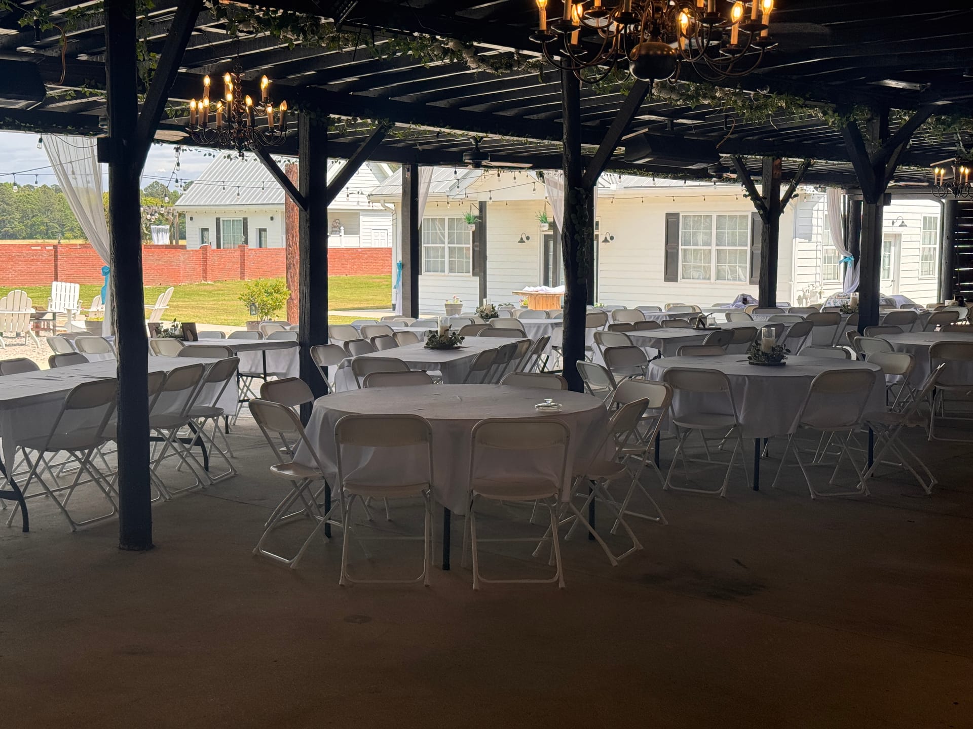 White-draped round reception tables and folding chairs arranged beneath a rustic open-air pavilion adorned with iron chandeliers, string lights, and green vine accents at Scarborough House wedding venue.