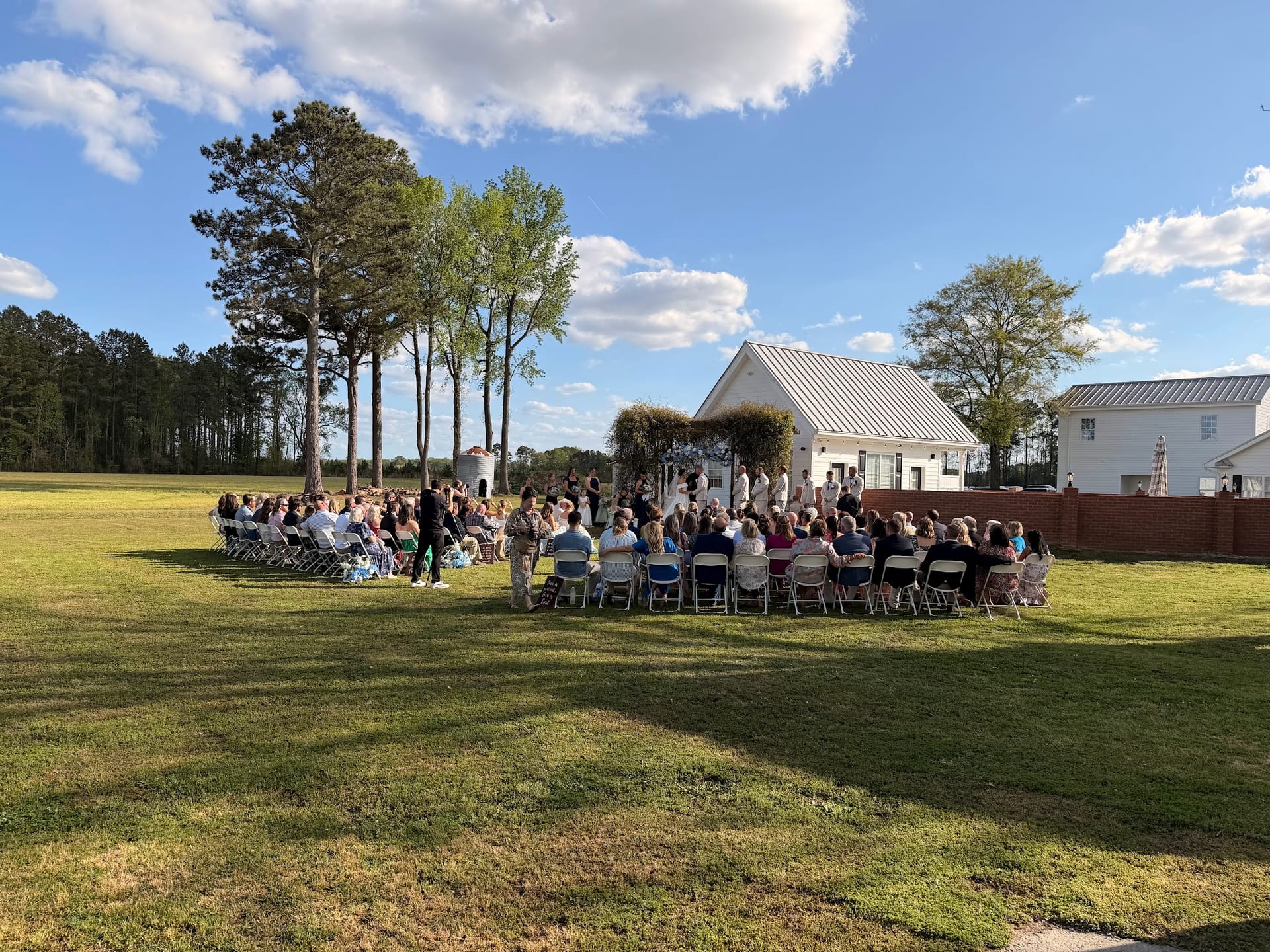 Guests seated in rows of white folding chairs on a sunlit lawn watch an outdoor wedding ceremony beneath a floral arch with a white manor house and tall trees in the background at Scarborough House.