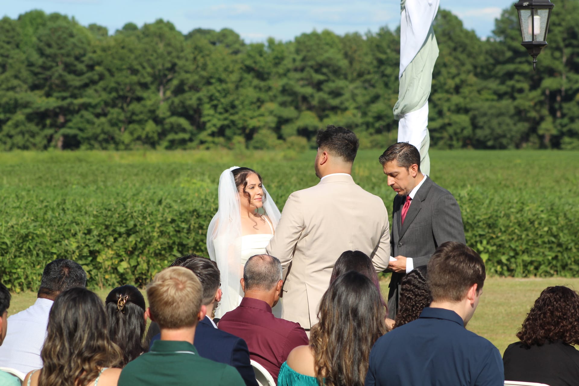 Bride in white gown and veil smiles at groom during outdoor wedding ceremony on the ceremony lawn at Scarborough House, with lush green farmland fields and trees in the background.