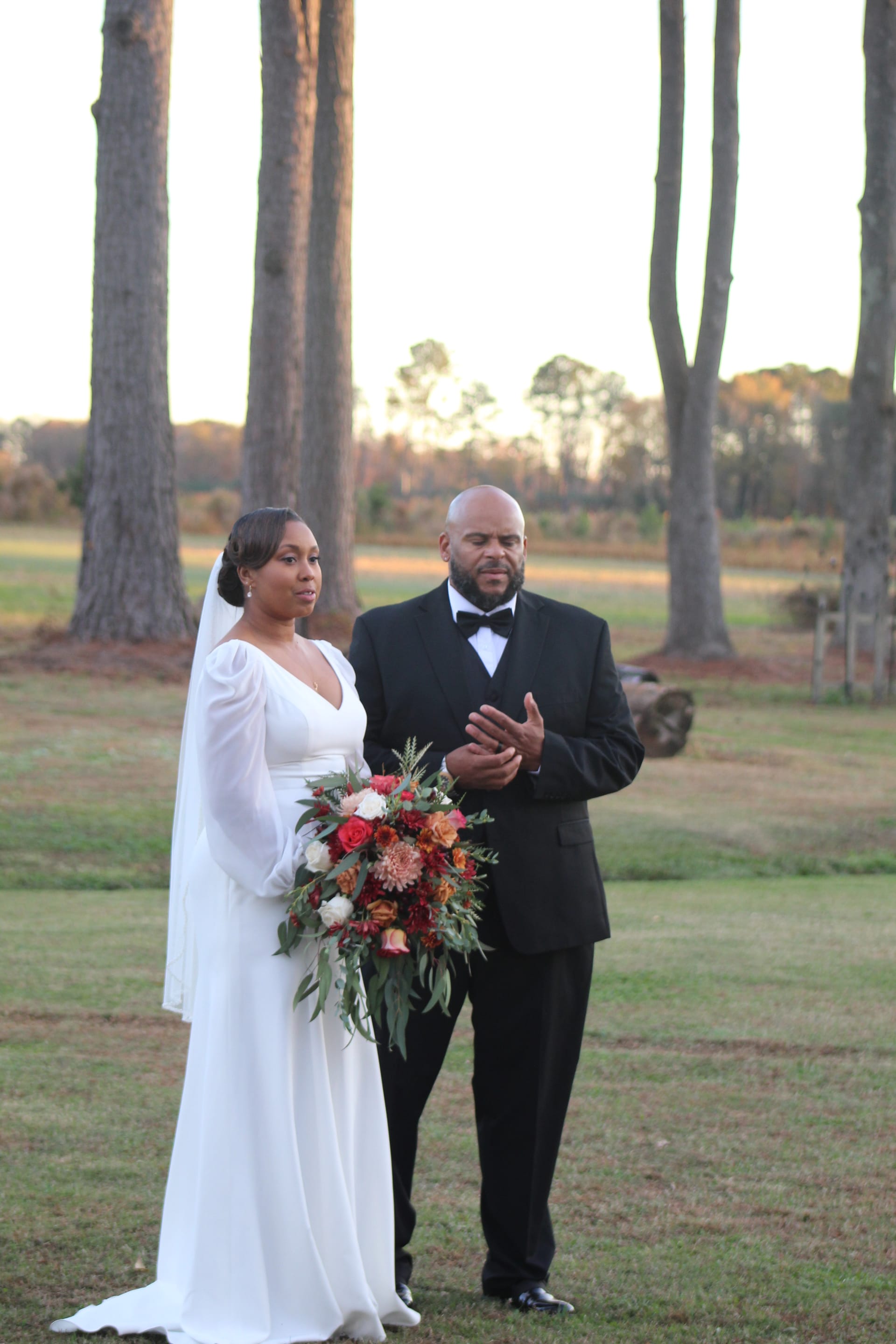 Bride in a long-sleeve white gown holding a lush autumn bouquet stands beside groom in a black tuxedo during an outdoor wedding ceremony on the historic Scarborough House ceremony lawn surrounded by tall pine trees at golden hour.