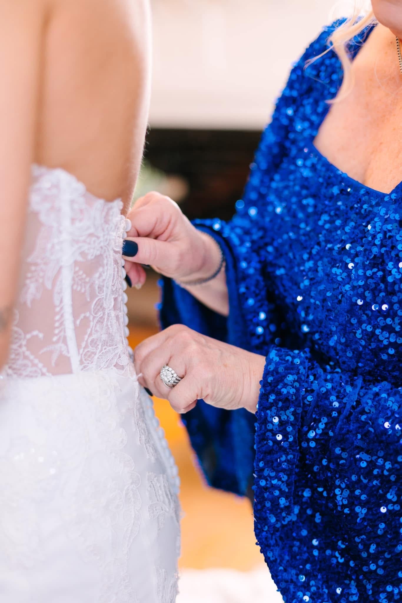 A woman in a royal blue sequined gown carefully fastens the lace back of a brides wedding dress while wearing a sparkling diamond ring.