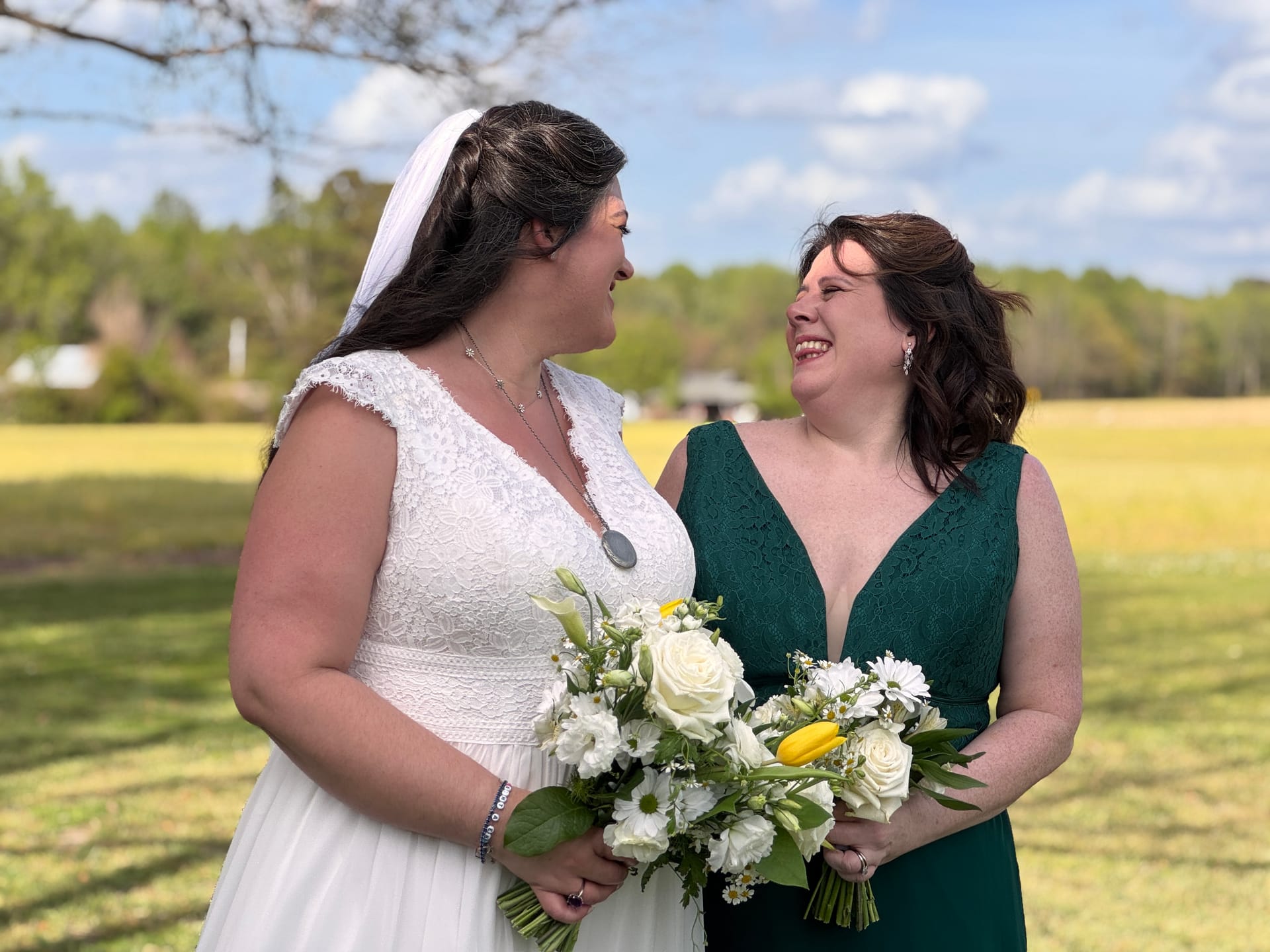Bride in white lace gown and veil shares a joyful laugh with her bridesmaid in an emerald green dress while holding white and yellow floral bouquets against a sunny farmland field at Scarborough House.