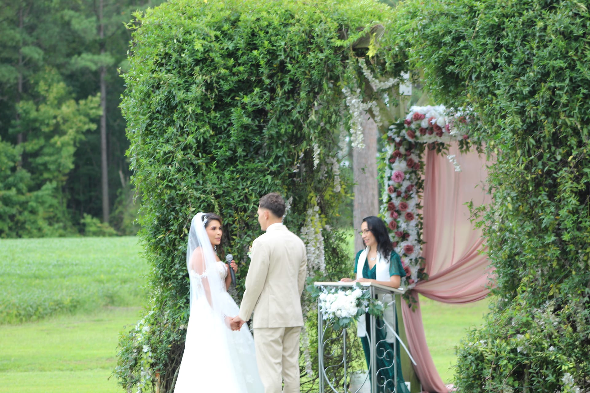 A bride and groom exchange vows beneath a lush ivy-covered archway adorned with pink drapery and floral arrangements while an officiant presides at Scarborough House estate.
