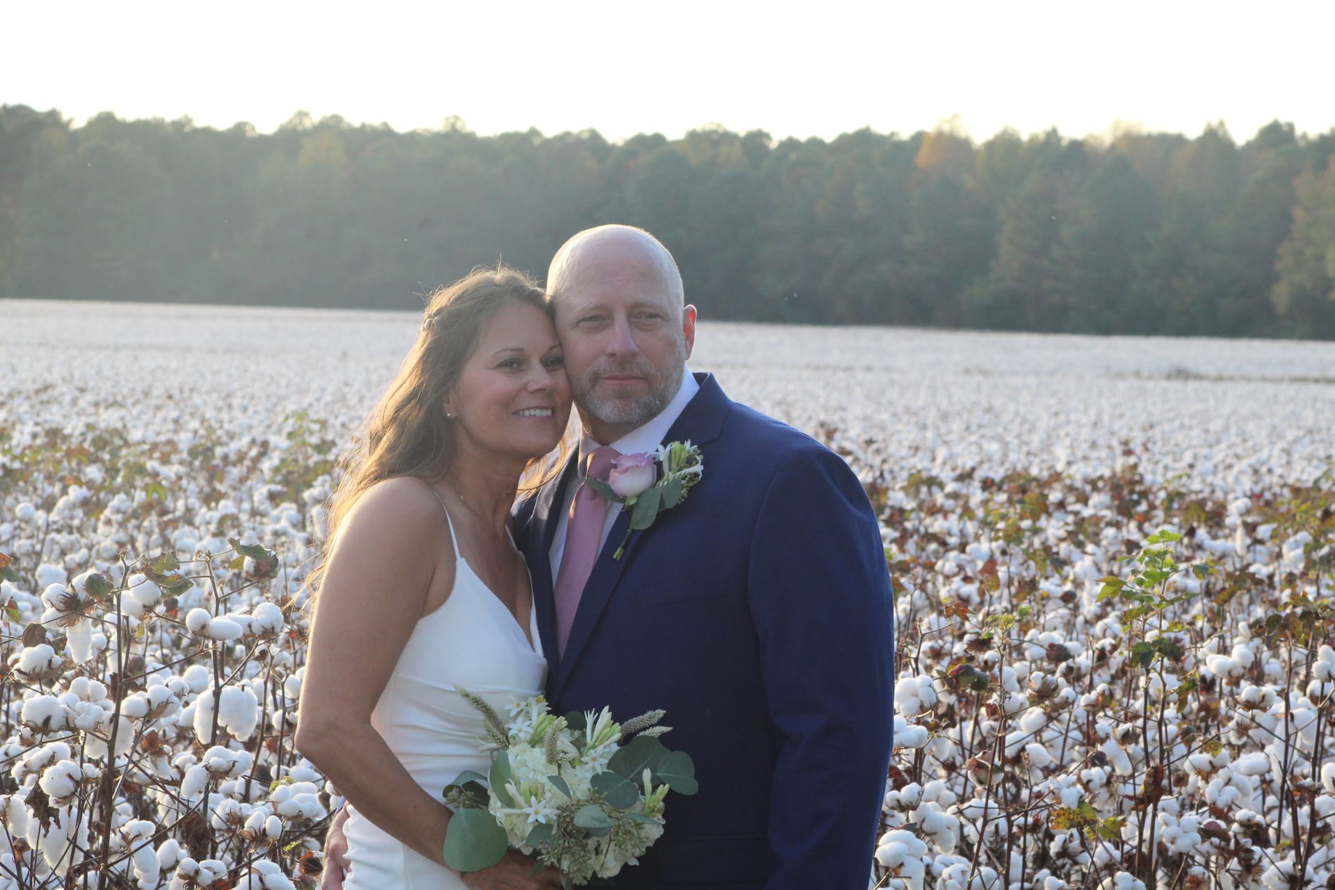 Bride in white gown holding a floral bouquet leans against groom in navy suit with pink tie and boutonniere as they pose together in a sunlit cotton field at Scarborough House.