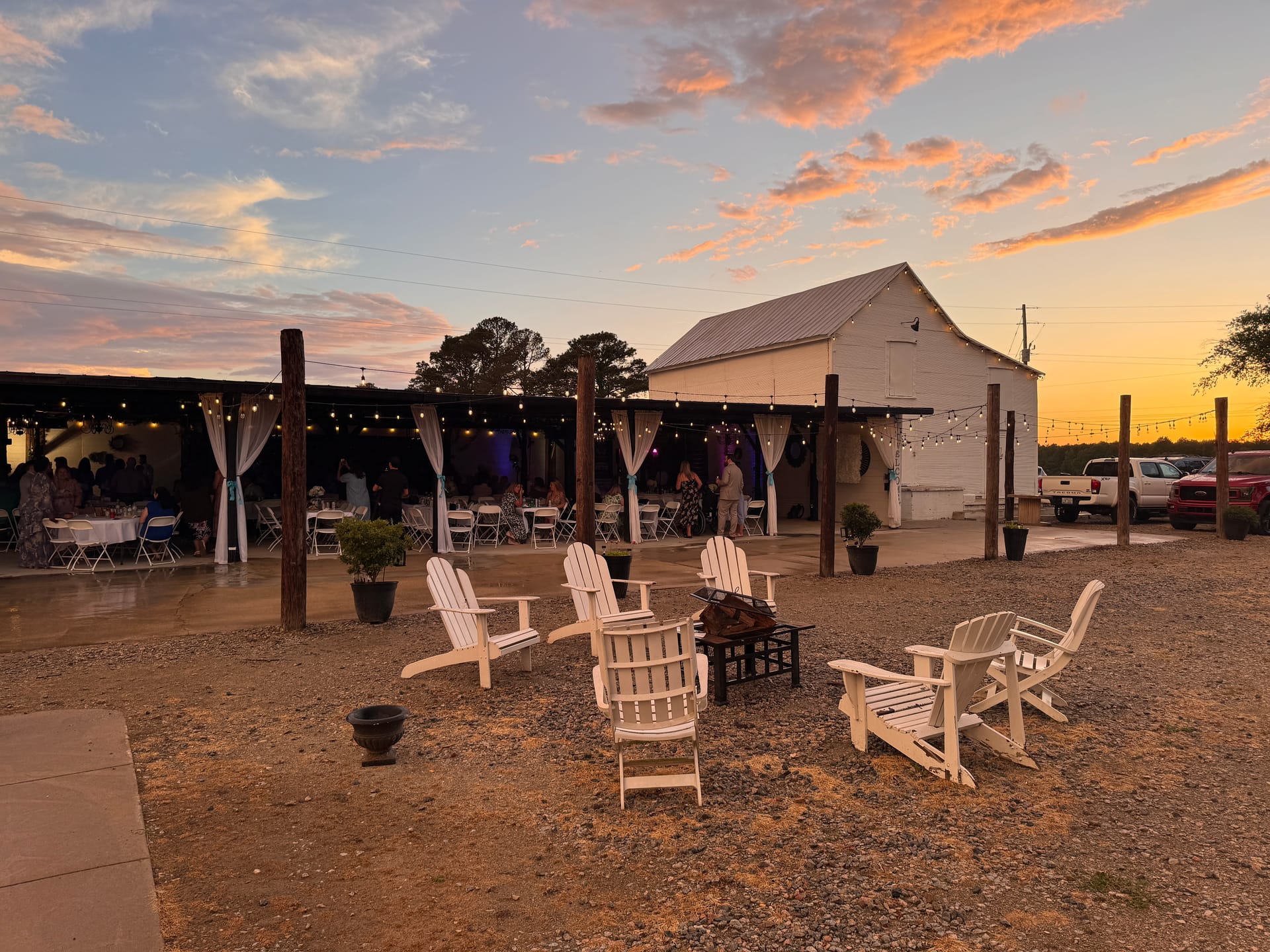 White Adirondack chairs around a fire pit at dusk with a rustic pavilion in the background under a vivid sunset sky.