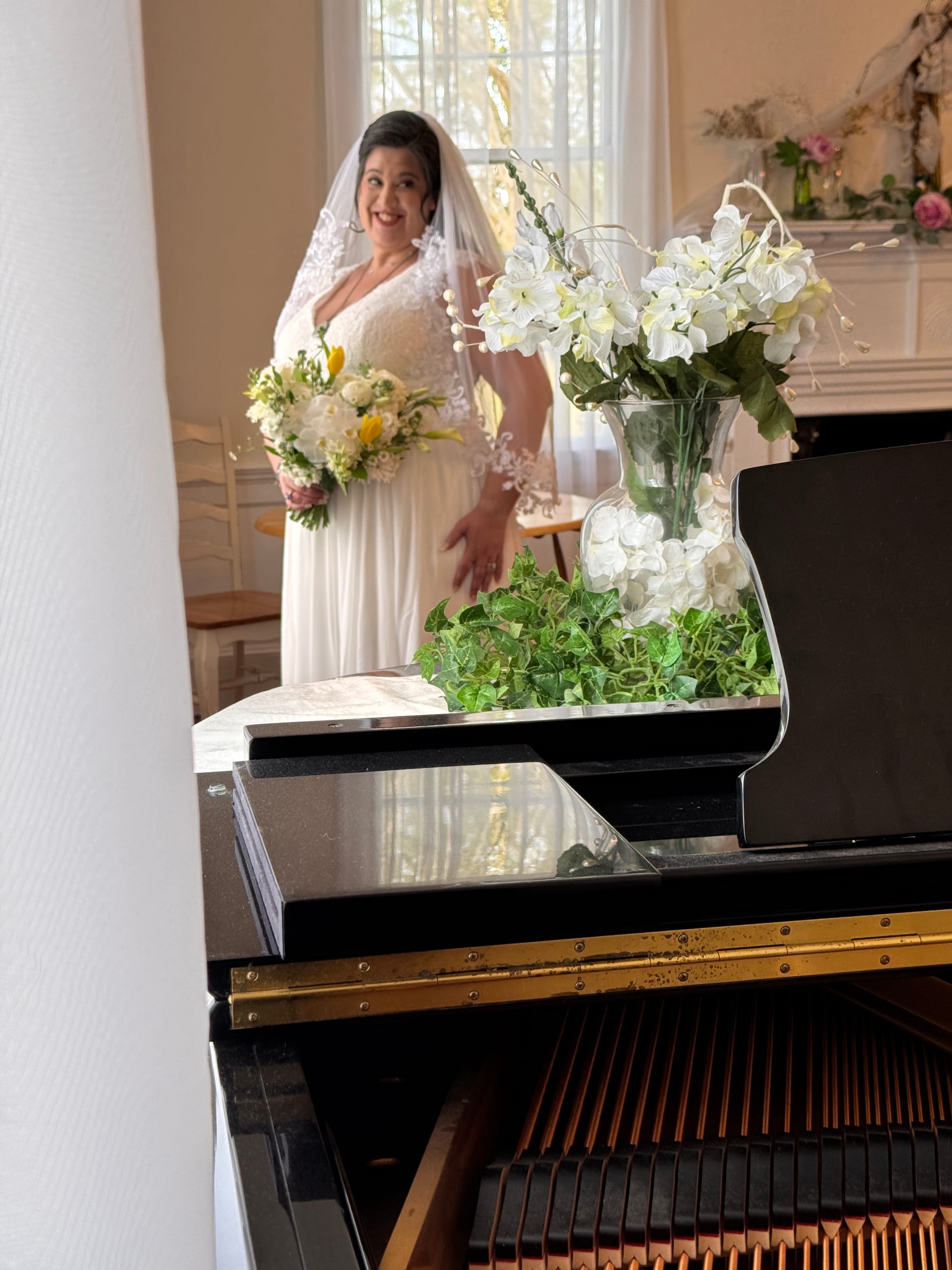 Smiling bride in a lace wedding gown and veil holds a bouquet of white and yellow flowers while standing near a window in the grand hall, with a black grand piano and white orchid arrangement in the foreground.