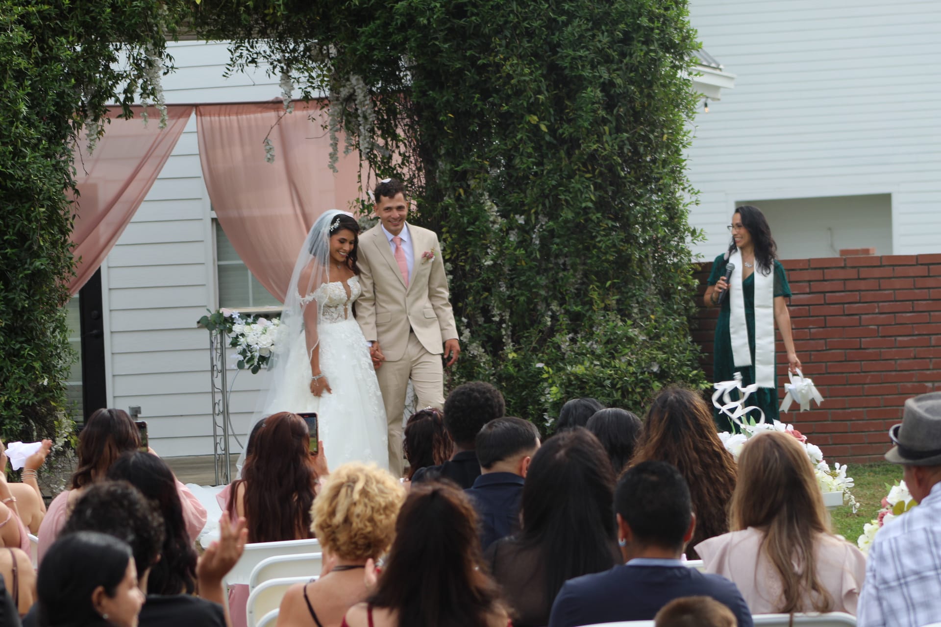 Smiling bride in a white lace gown and groom in a tan suit walk hand-in-hand down the aisle during their outdoor wedding recessional at Scarborough House, framed by a blush pink draped arch and lush greenery.