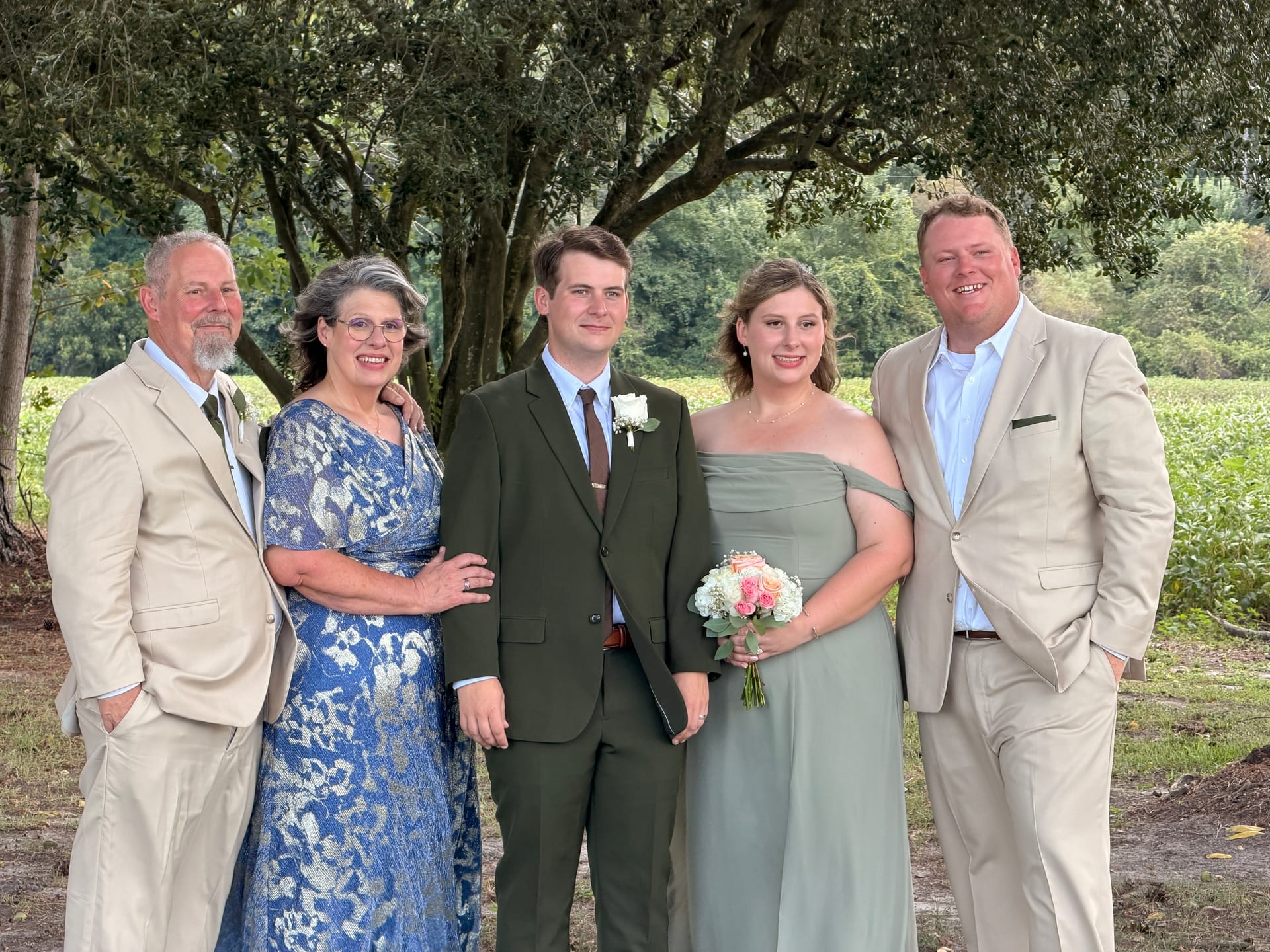 A smiling family of five poses outdoors beneath oak trees at Scarborough House estate, with the groom in a dark green suit and boutonniere and a bridesmaid holding a pink and white bouquet.